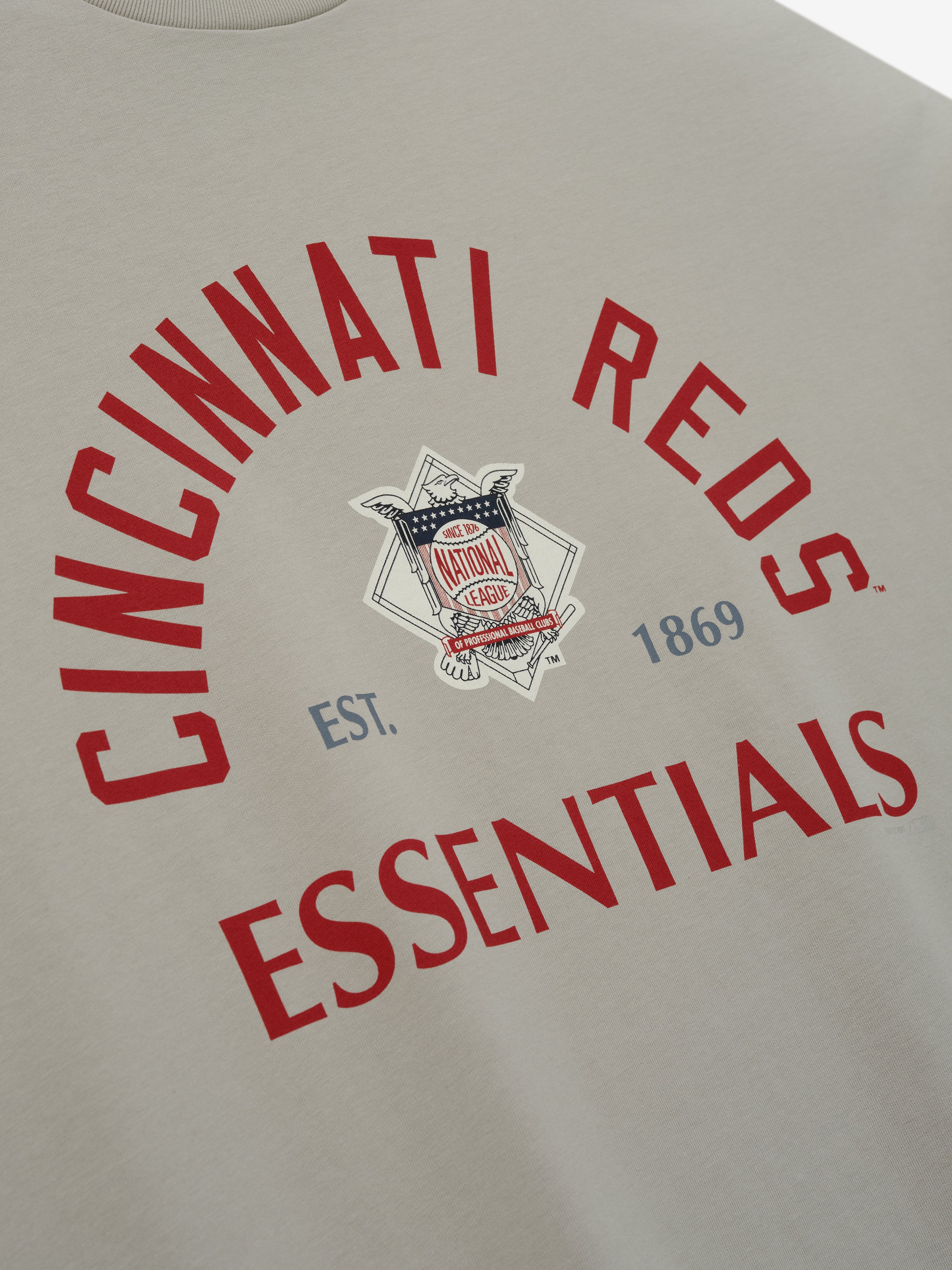 Reds NL Merch Tee in soft beige fabric featuring bold red arched "Cincinnati Reds" text and "Essentials" below, with a vintage-inspired emblem and establishment date in muted blue.