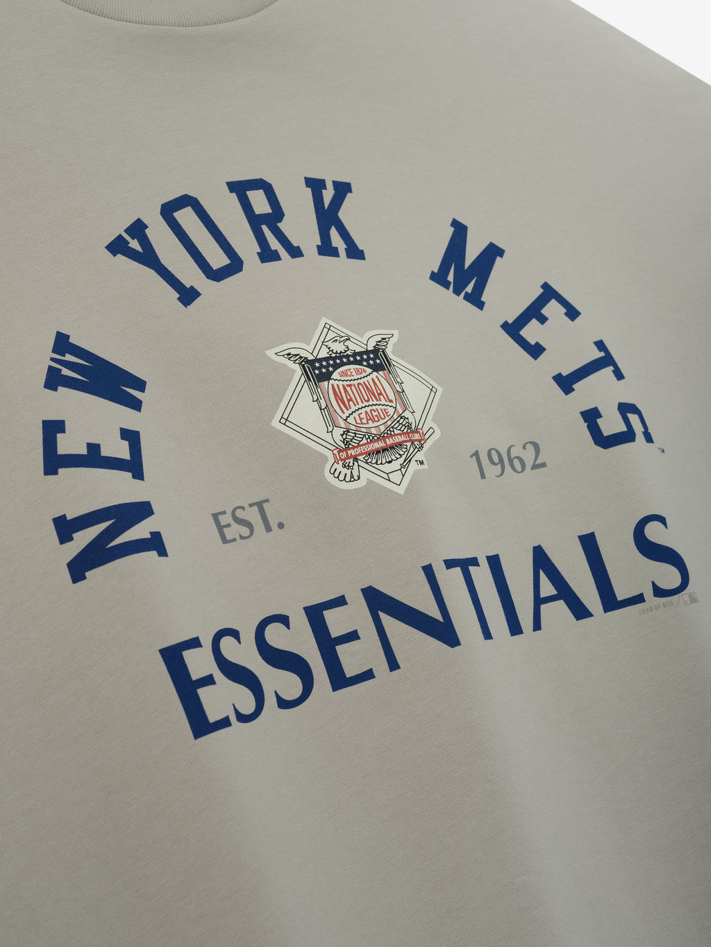 Close-up of the Mets NL Merch Tee in a muted gray tone featuring bold navy blue text reading "New York Mets" arching above a vintage-style National League emblem with "ESSENTIALS" printed below.
