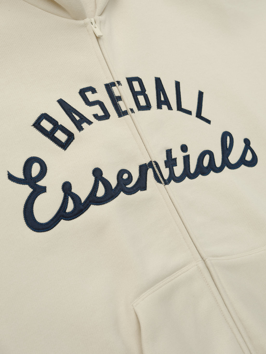 Close-up of cream-colored Mlb Full Zip Sport Hoodie featuring navy embroidered "Baseball Essentials" text above the front kangaroo pockets with a full zipper closure.