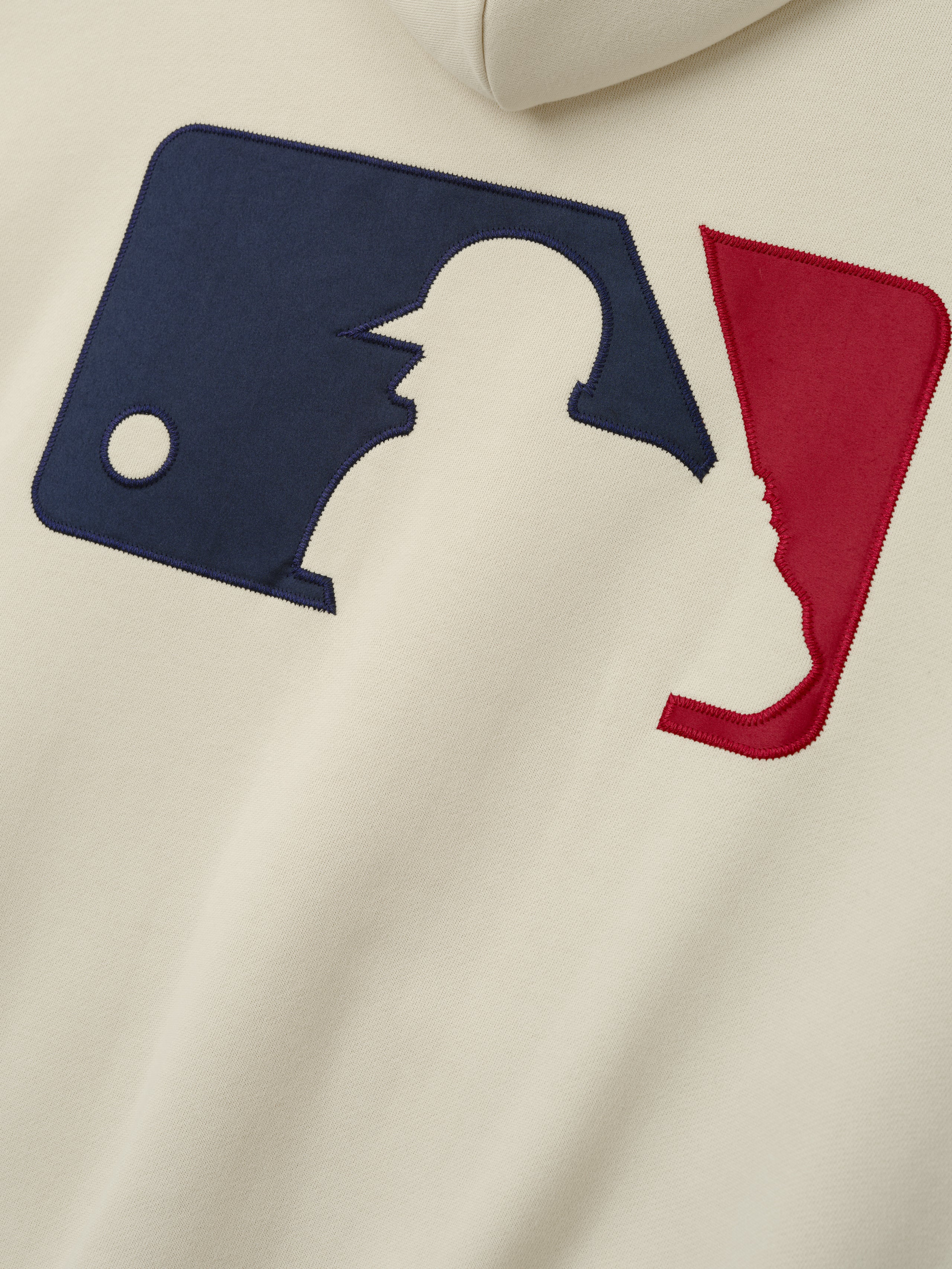 Close-up of the back of an off-white Mlb Full Zip Sport Hoodie featuring a large stitched baseball logo in blue and red fabric with clean, detailed embroidery.