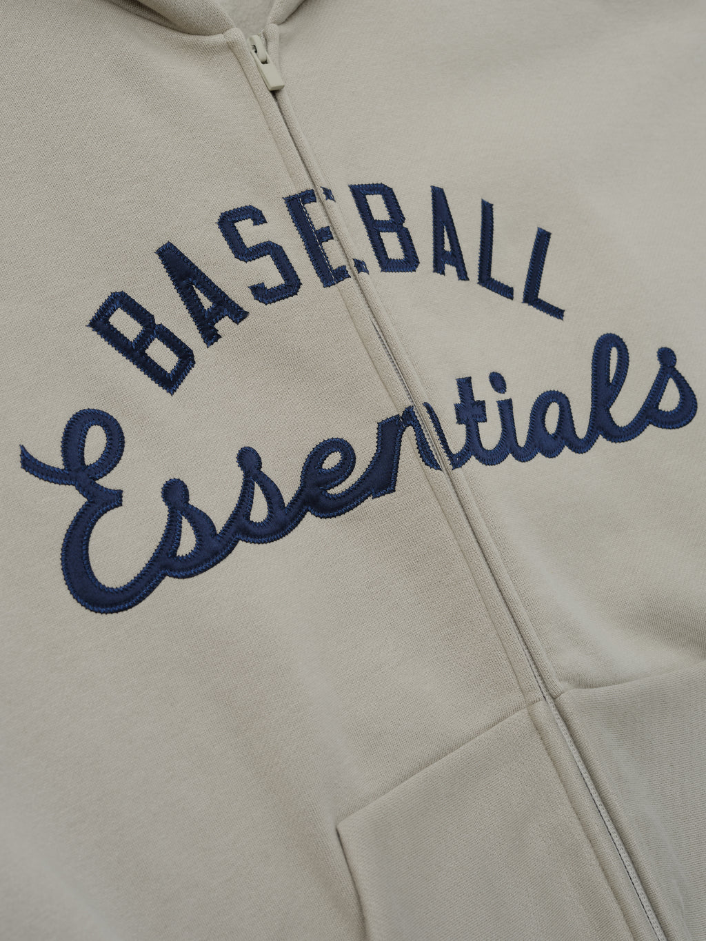 Close-up of a muted taupe Mlb Full Zip Sport Hoodie highlighting navy embroidered "Baseball Essentials" text across the front with a full front zipper and kangaroo pocket.