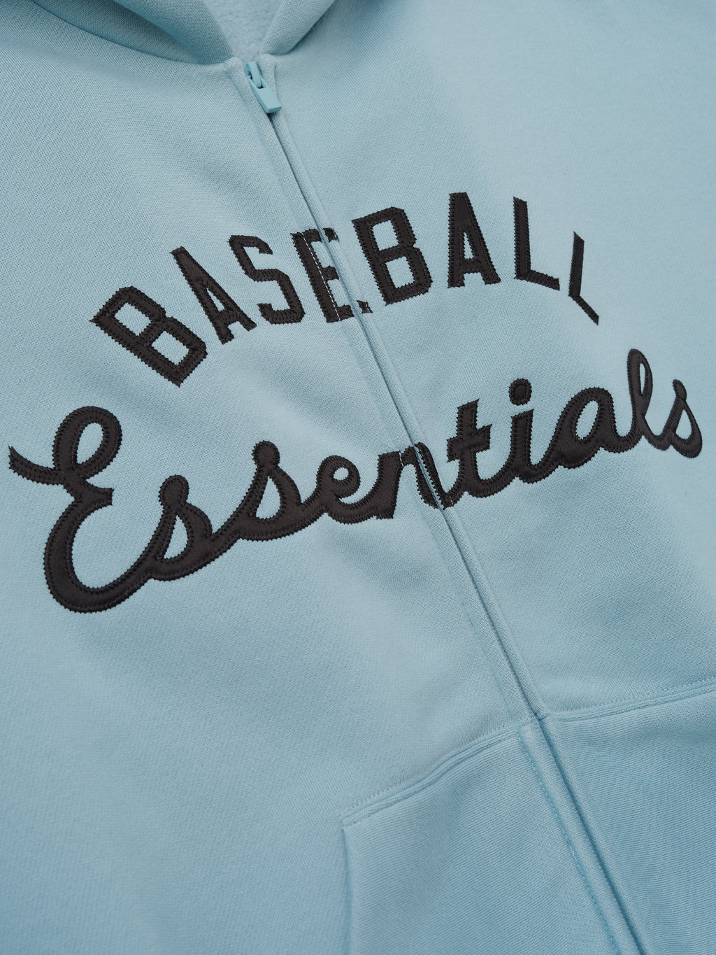 Close-up of Mlb Full Zip Sport Hoodie in soft stone blue showing black embroidered "Baseball Essentials" text above the front kangaroo pocket with visible zipper and hood fabric.