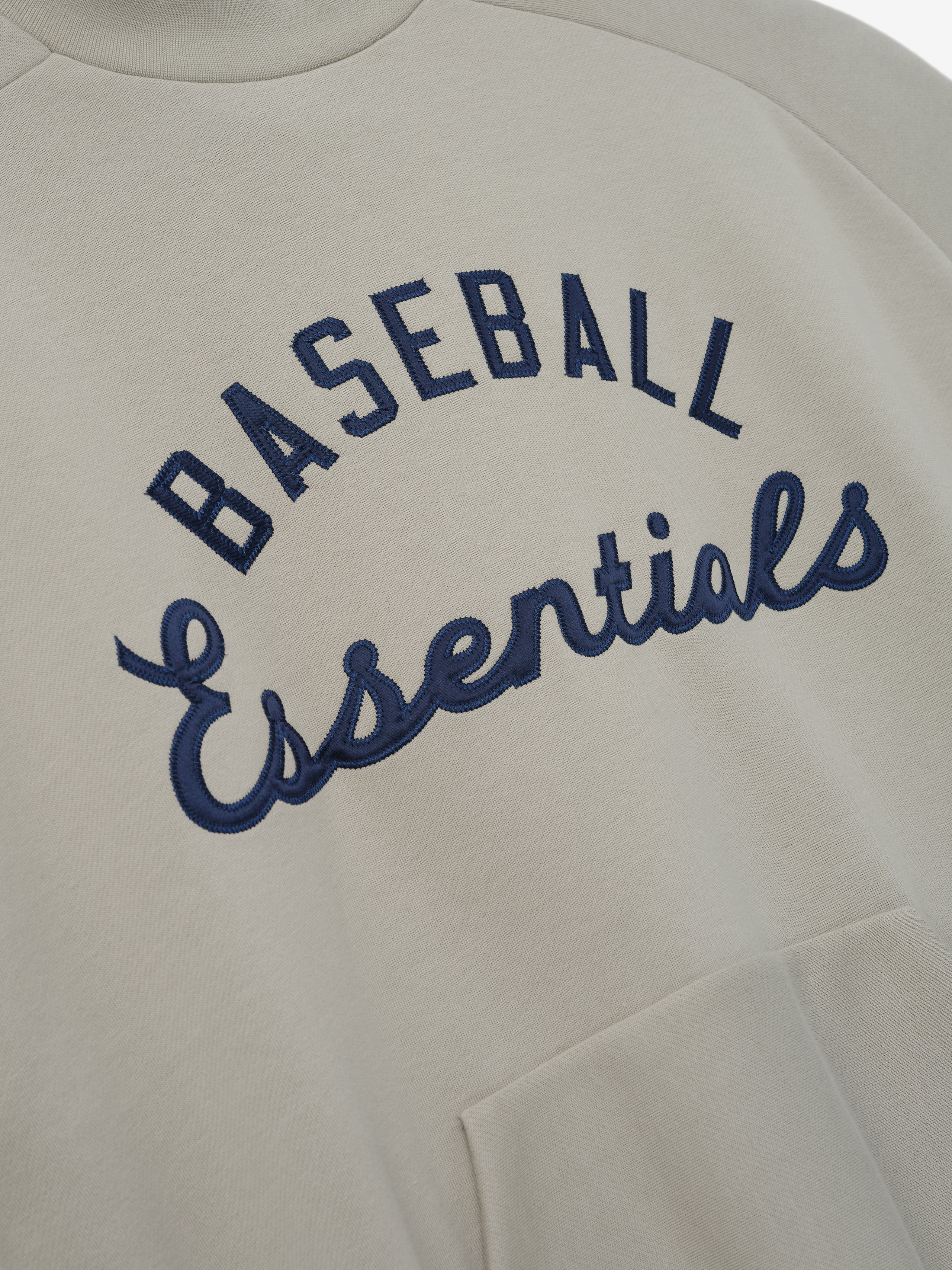 Mlb Sport Crewneck in a muted beige tone featuring navy blue embroidered "Baseball Essentials" text arching across the chest with a soft fabric and a front kangaroo pocket for casual layering.