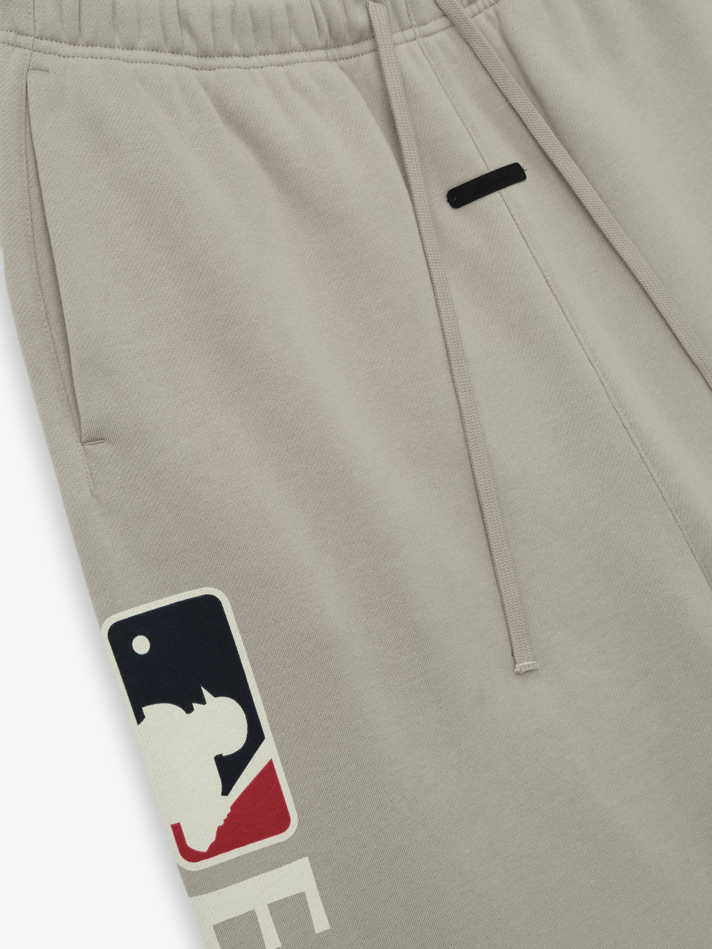 Close-up of Mlb Lounge Sweatpant in soft beige fabric featuring an elastic waistband with drawstrings and a bold baseball-themed graphic near the side pocket on casual bottoms.