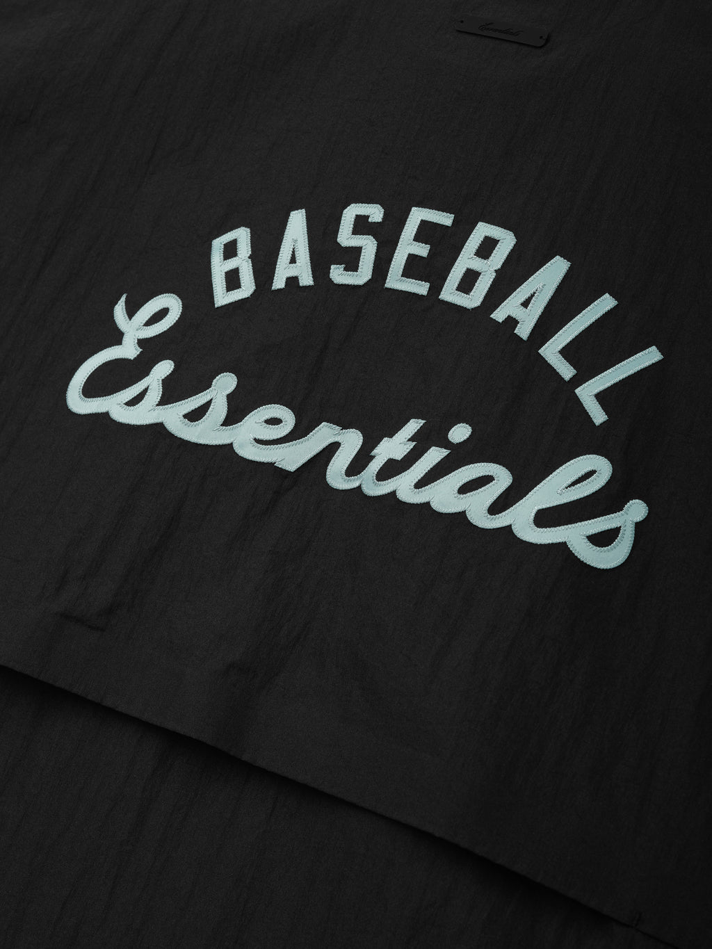 Close-up of black fabric from the Mlb 3/4 Sleeve Pullover featuring raised light blue "BASEBALL Essentials" text in contrasting fonts on a smooth, lightweight material.