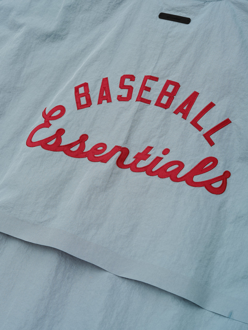 Close-up of the back panel on the Mlb 3/4 Sleeve Pullover in muted stone blue, featuring bold red "Baseball Essentials" text embroidered on lightweight fabric.