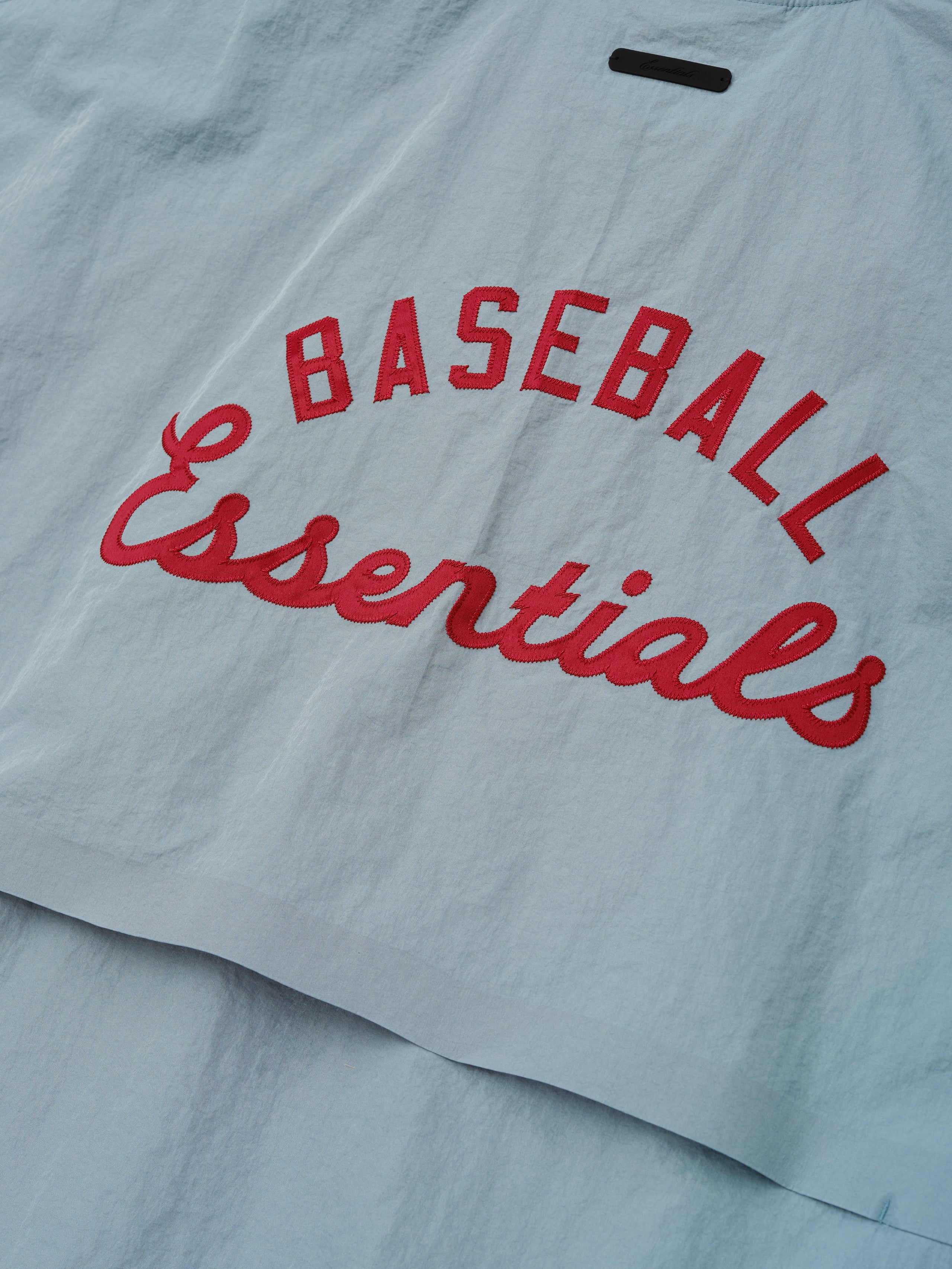 Close-up of the back panel on the Mlb 3/4 Sleeve Pullover in muted stone blue, featuring bold red "Baseball Essentials" text embroidered on lightweight fabric.