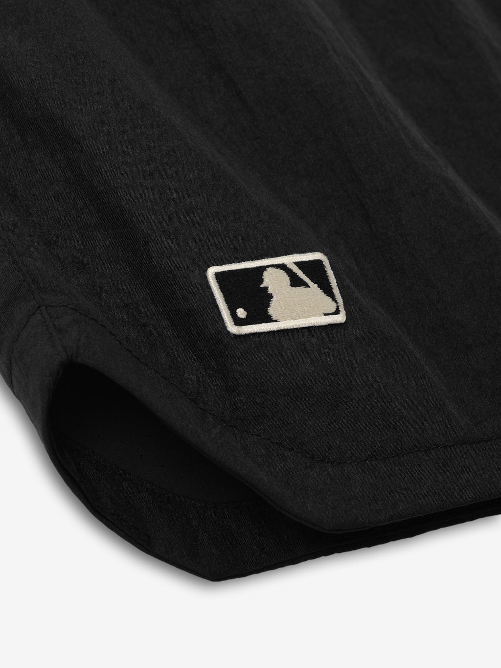 Close-up of the bottom hem of Mlb Gym Short in black fabric featuring a small embroidered baseball player logo near the side seam on lightweight athletic bottoms.