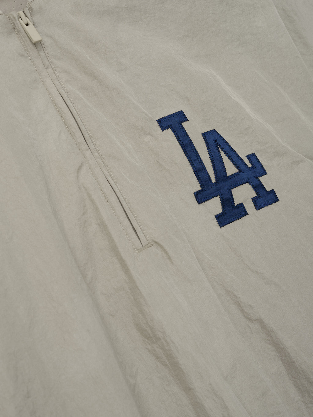 Close-up of a light beige Dodger 3/4 Sleeve Pullover fabric featuring a navy blue LA logo embroidery and a vertical zipper pocket detail on smooth material.