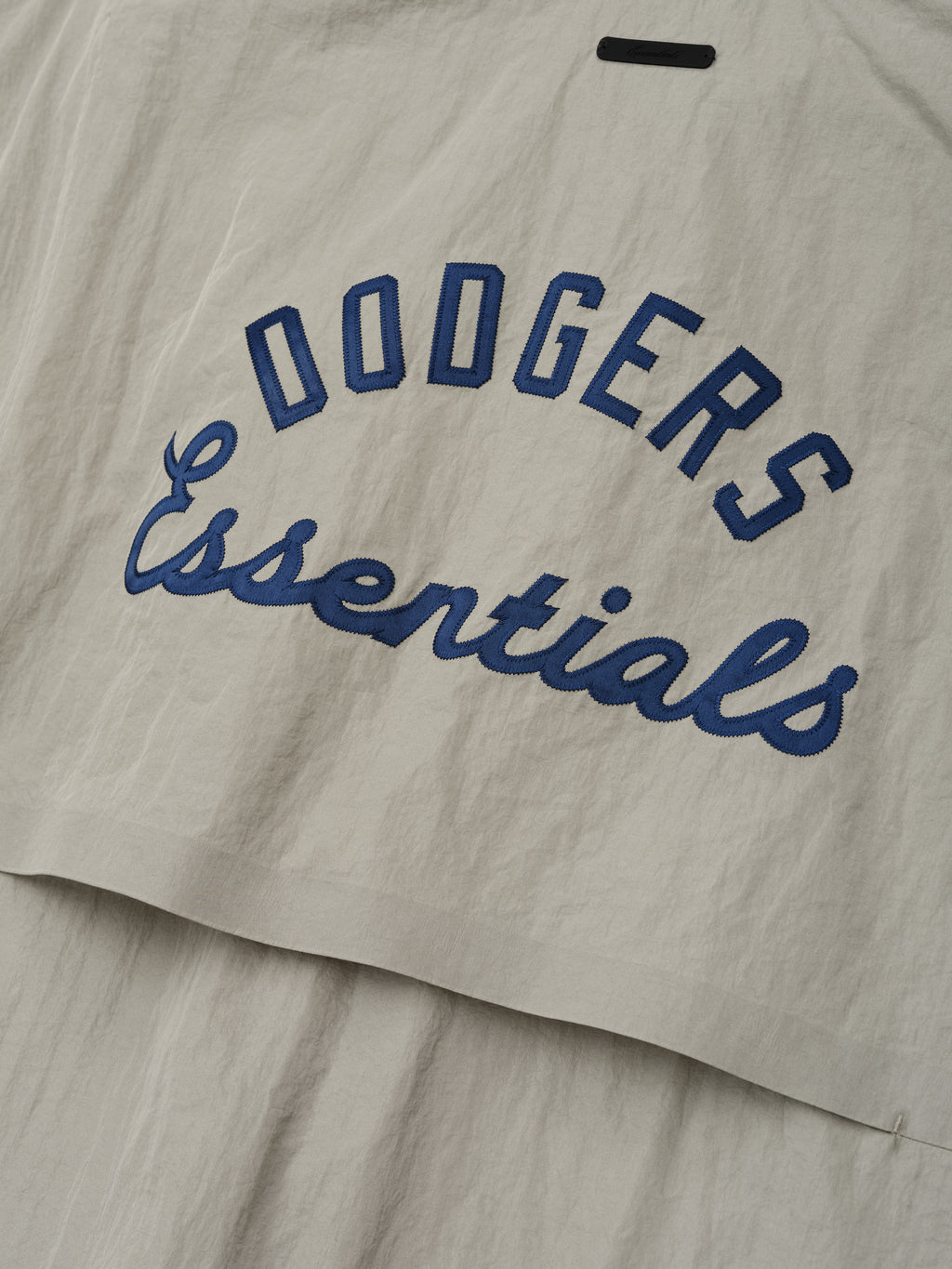 Close-up of a light beige Dodger 3/4 Sleeve Pullover fabric featuring navy blue "Dodgers Essentials" embroidered text in an arched and cursive style on a textured material.