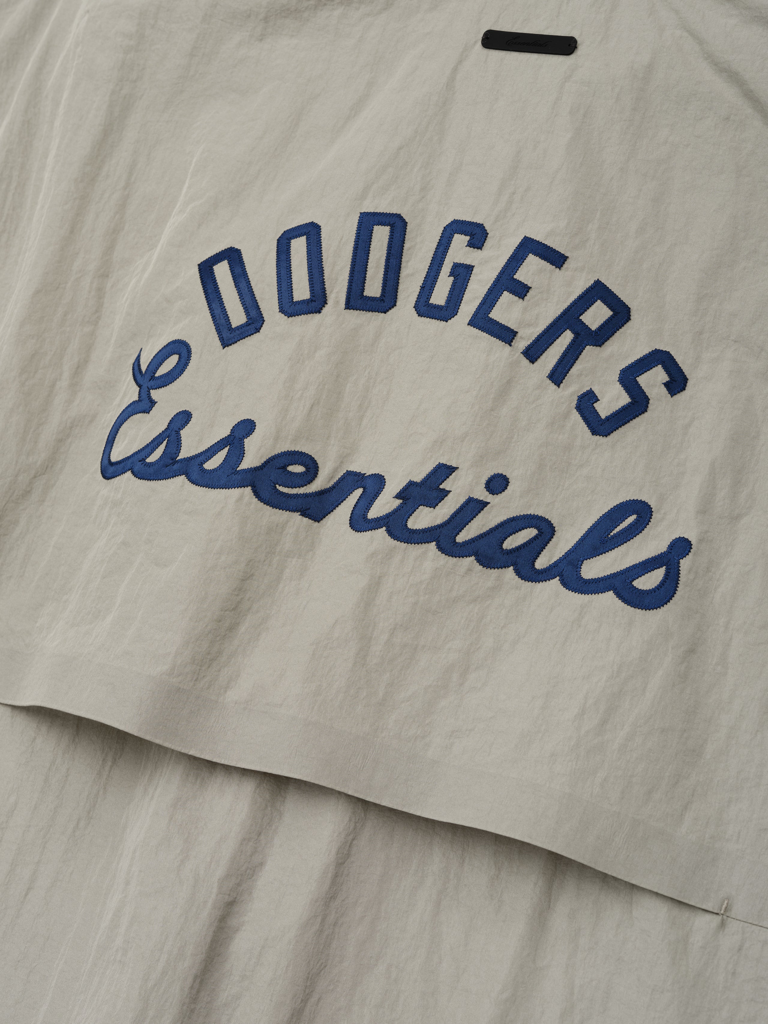 Close-up of a light beige Dodger 3/4 Sleeve Pullover fabric featuring navy blue "Dodgers Essentials" embroidered text in an arched and cursive style on a textured material.