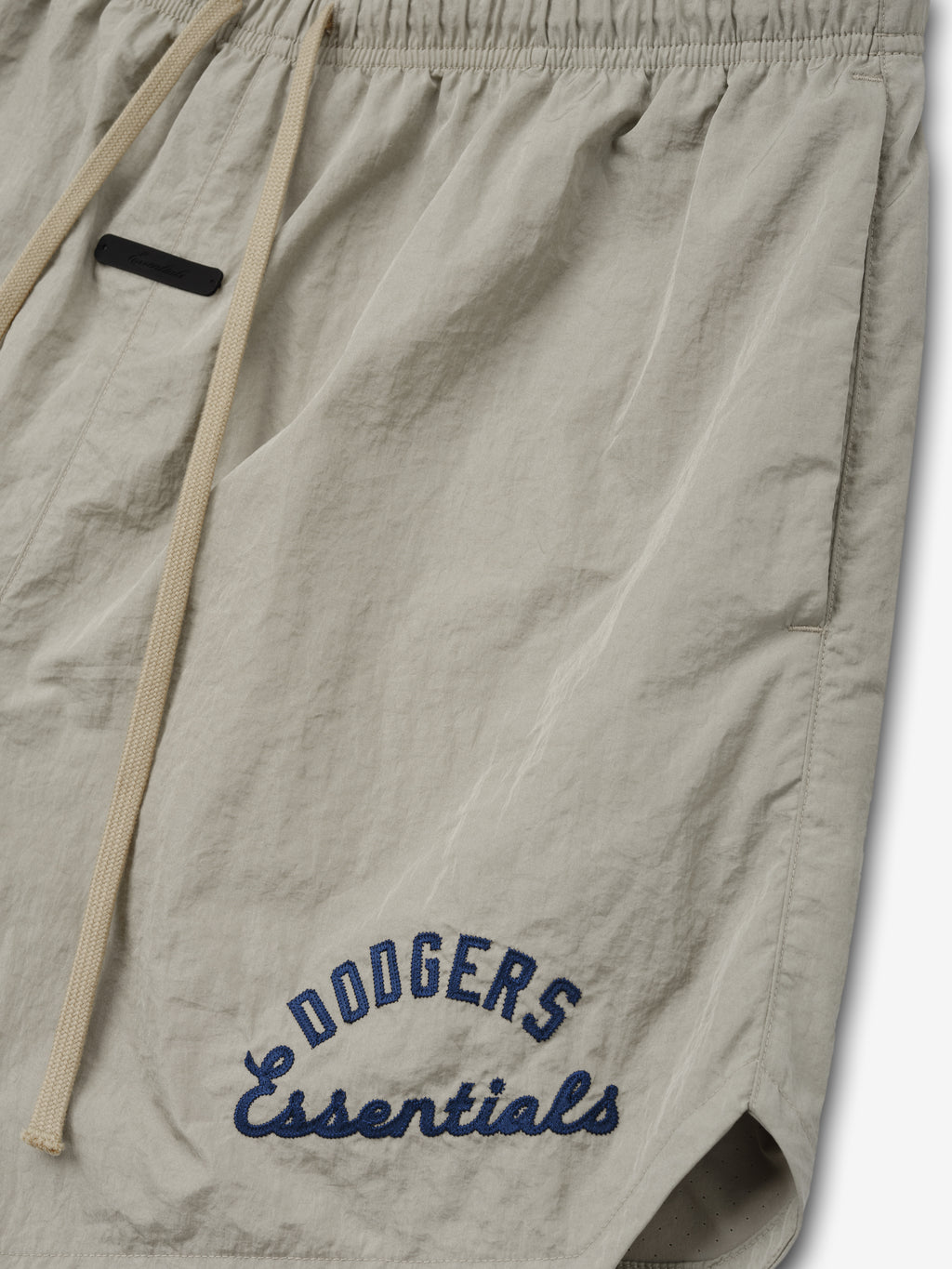 Close-up of light beige Dodgers Gym Short featuring navy blue embroidered "Dodgers Essentials" text on lightweight fabric with an elastic waistband and beige drawstrings.