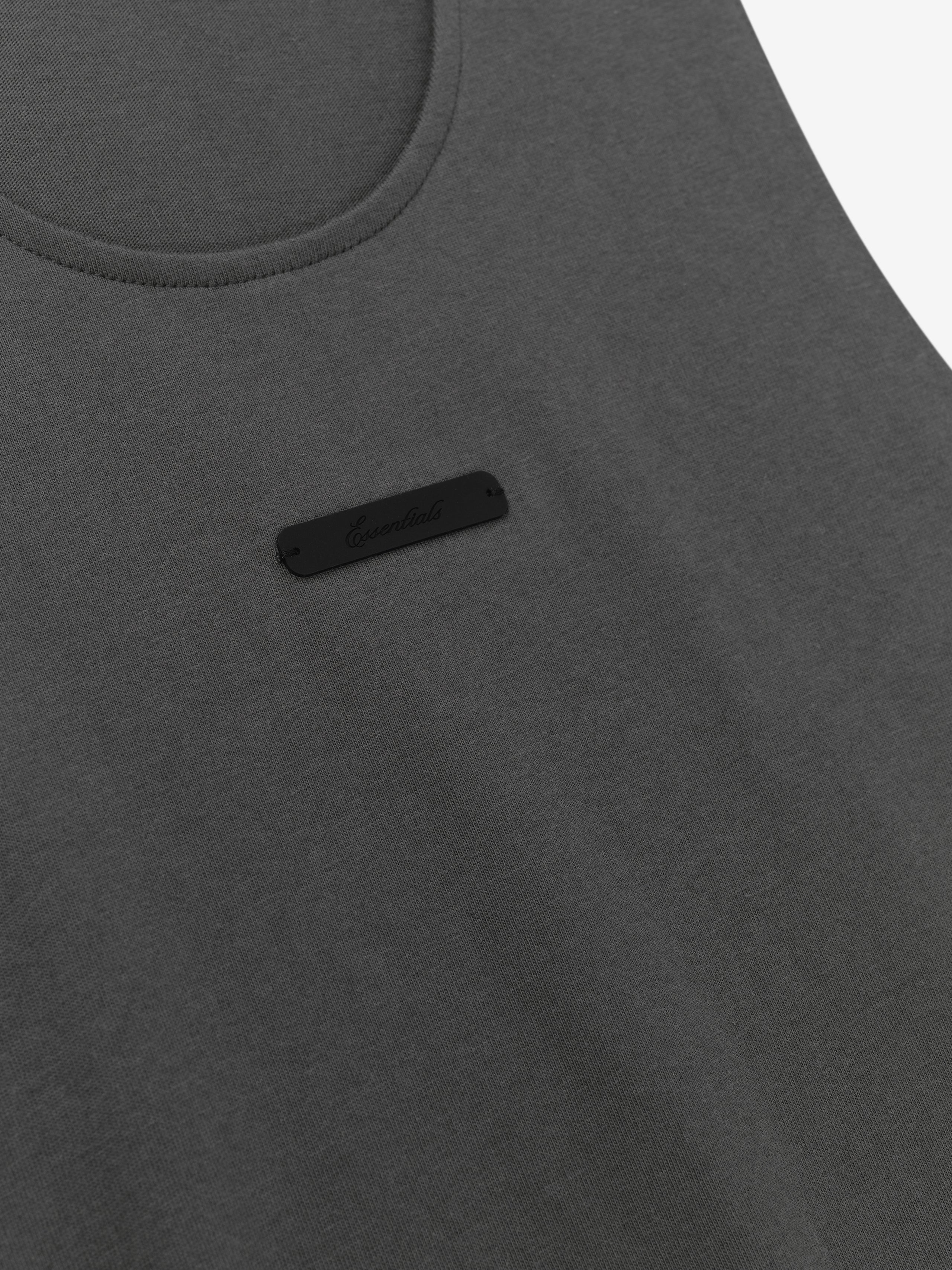 Essential Tank in a dark iron grey shade featuring a soft fabric with a rounded neckline and a subtle black logo patch on the chest for a simple, versatile top.