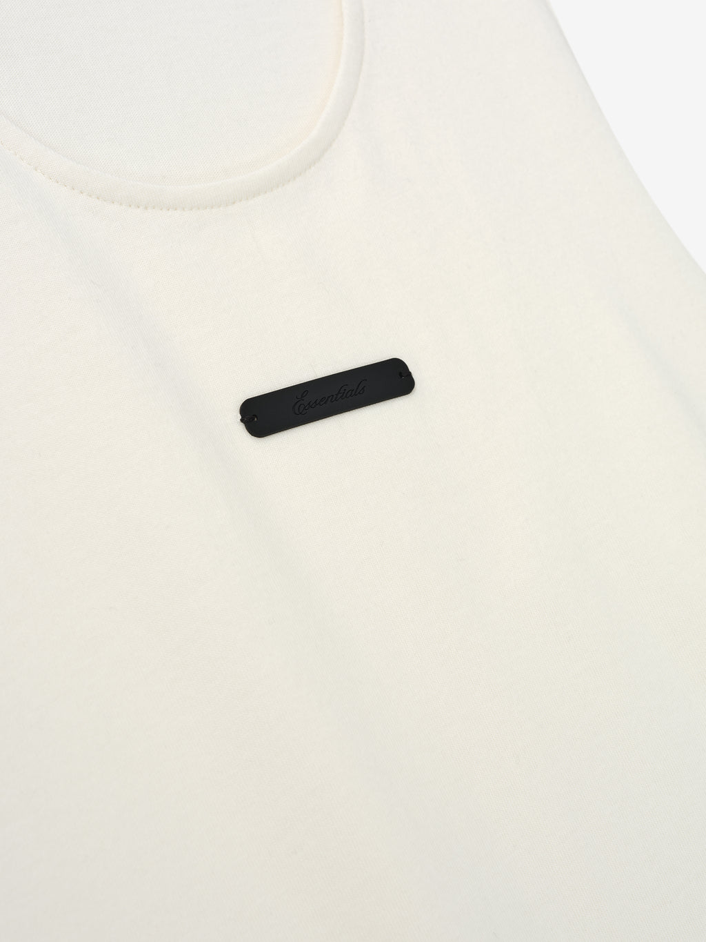 Essential Tank in a soft ivory hue featuring a smooth fabric with a rounded neckline and a small black rectangular logo patch centered on the chest for a refined casual top.