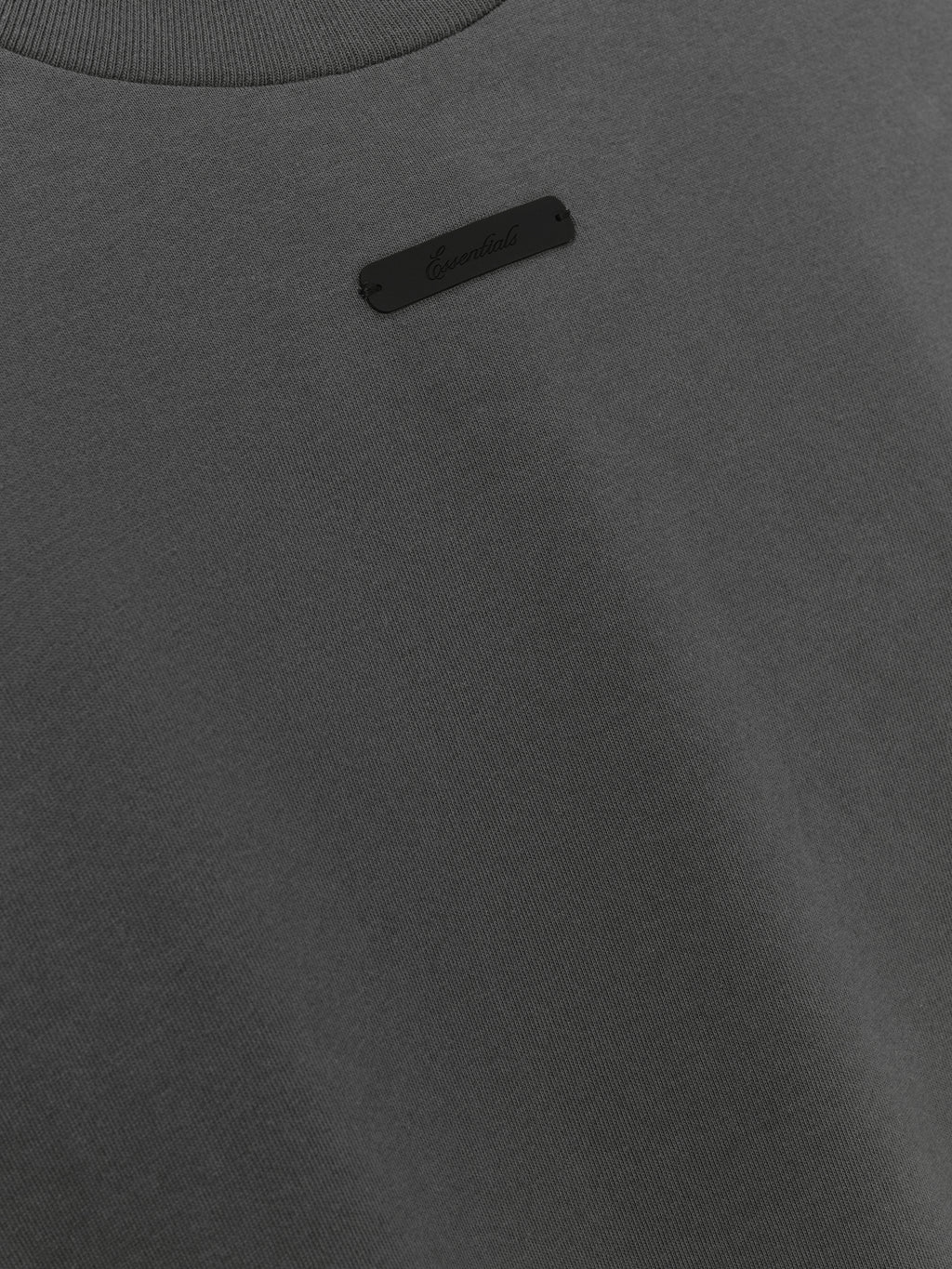 Merch Tee (Boxy) in dark grey featuring a minimalist design with a small black "Essentials" logo patch near the collar for a clean, understated casual top.