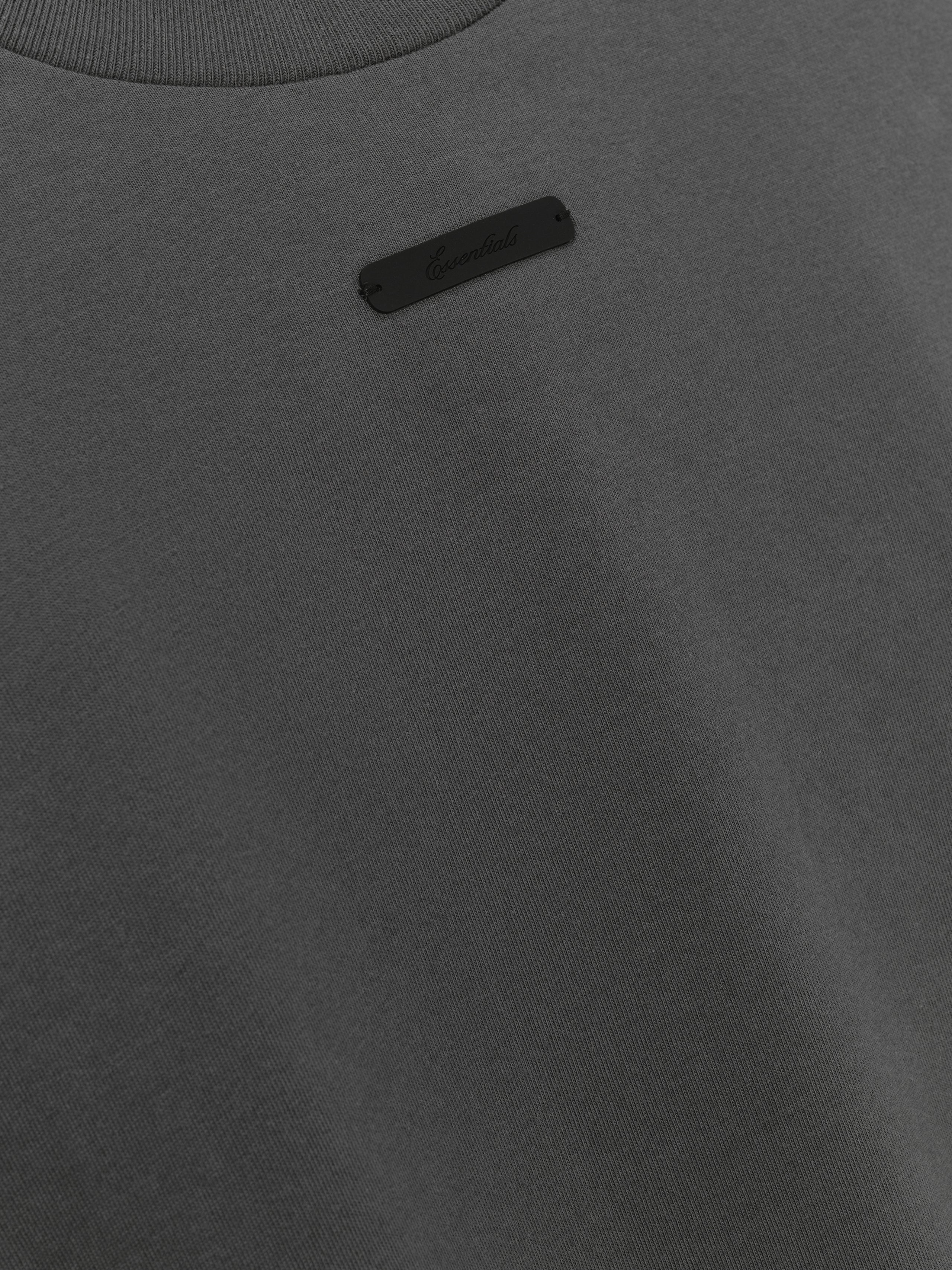 Merch Tee (Boxy) in dark grey featuring a minimalist design with a small black "Essentials" logo patch near the collar for a clean, understated casual top.