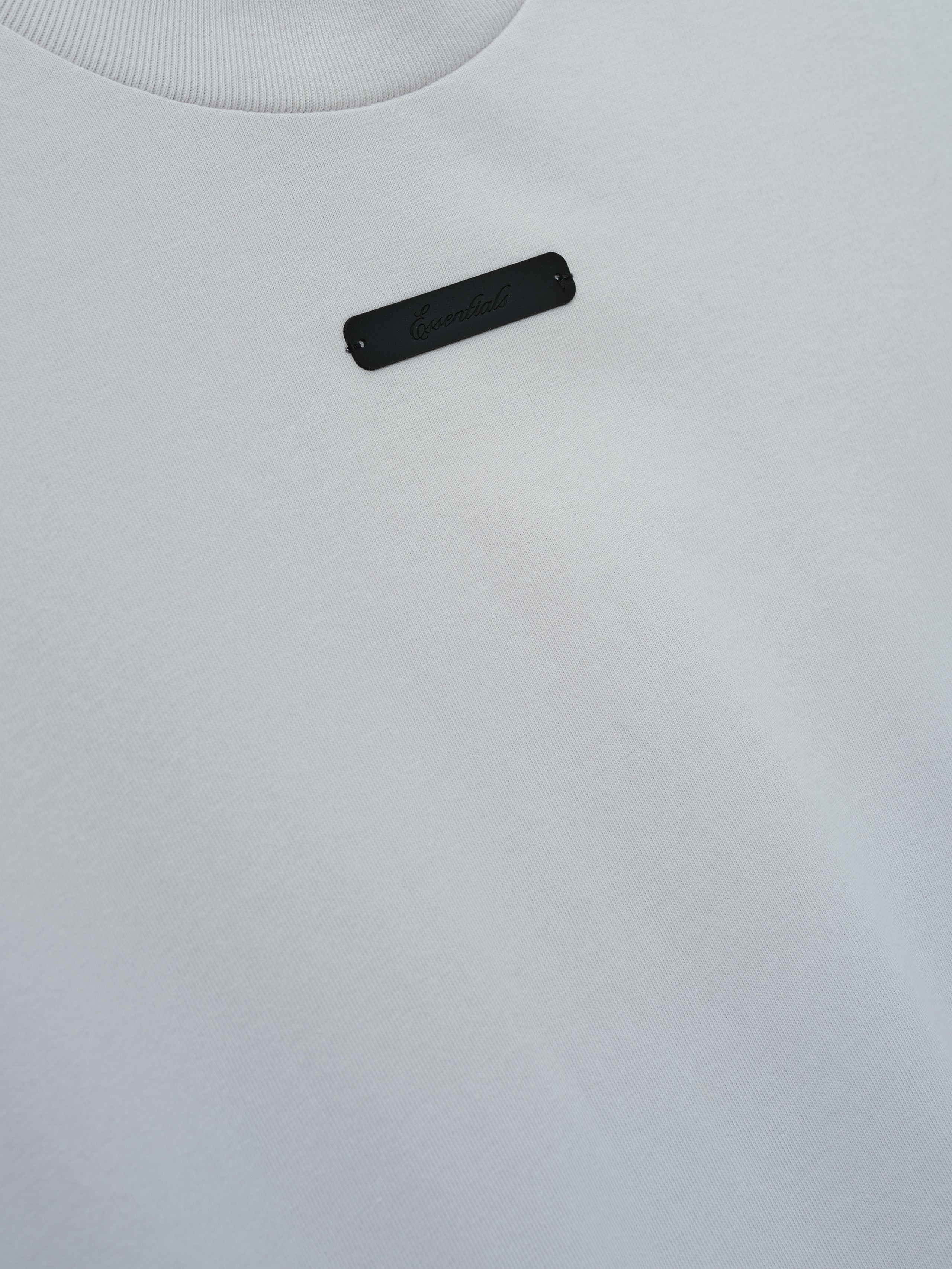 Merch Tee (Boxy) in soft light grey fabric featuring a minimalist design with a small subtle black rectangular detail on the upper chest for a clean and modern top.
