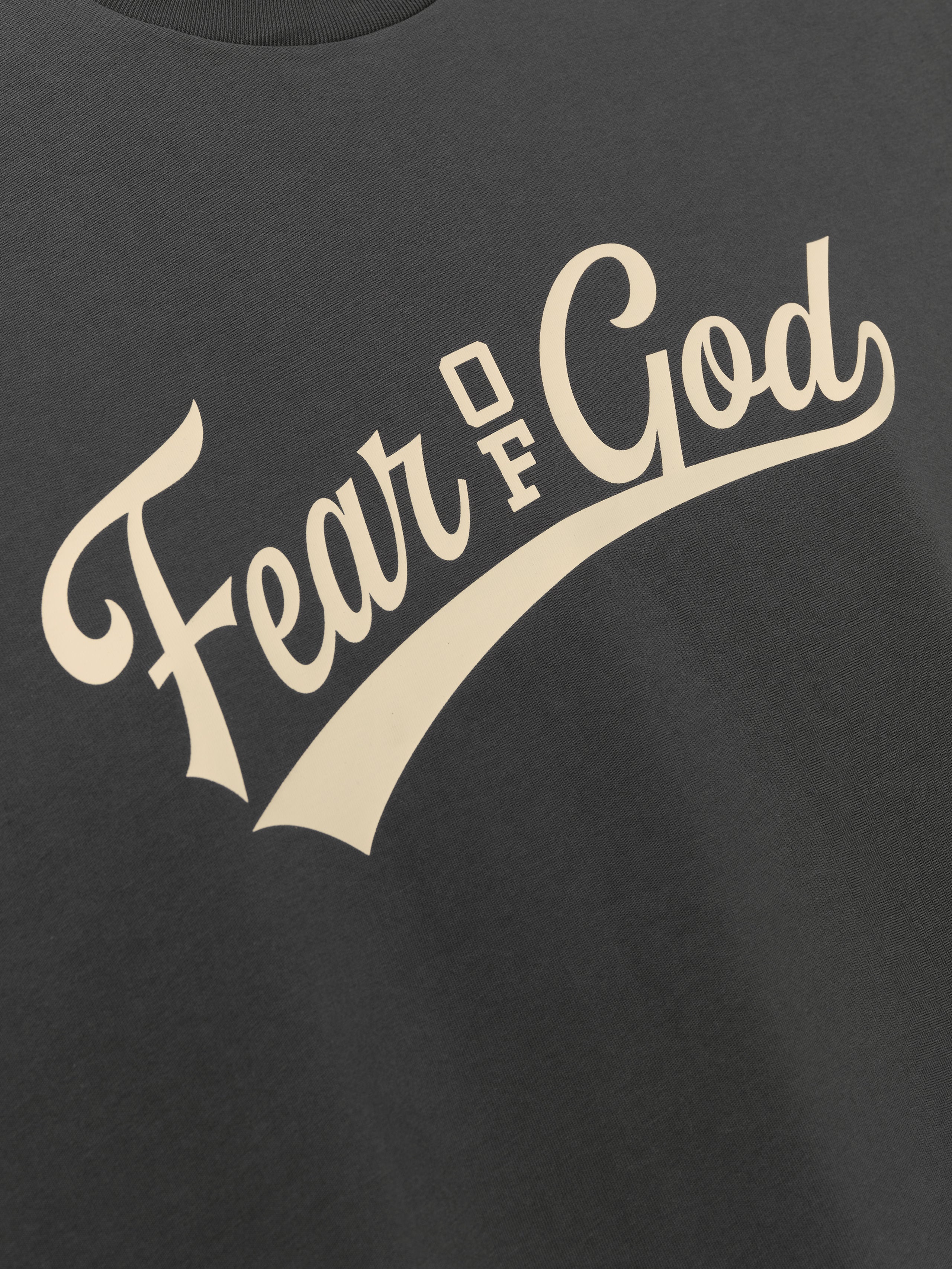 Merch Tee (Boxy) in dark iron grey featuring a relaxed fit with subtle texture and bold cream Fear of God script printed diagonally across the front for a vintage-inspired casual top.