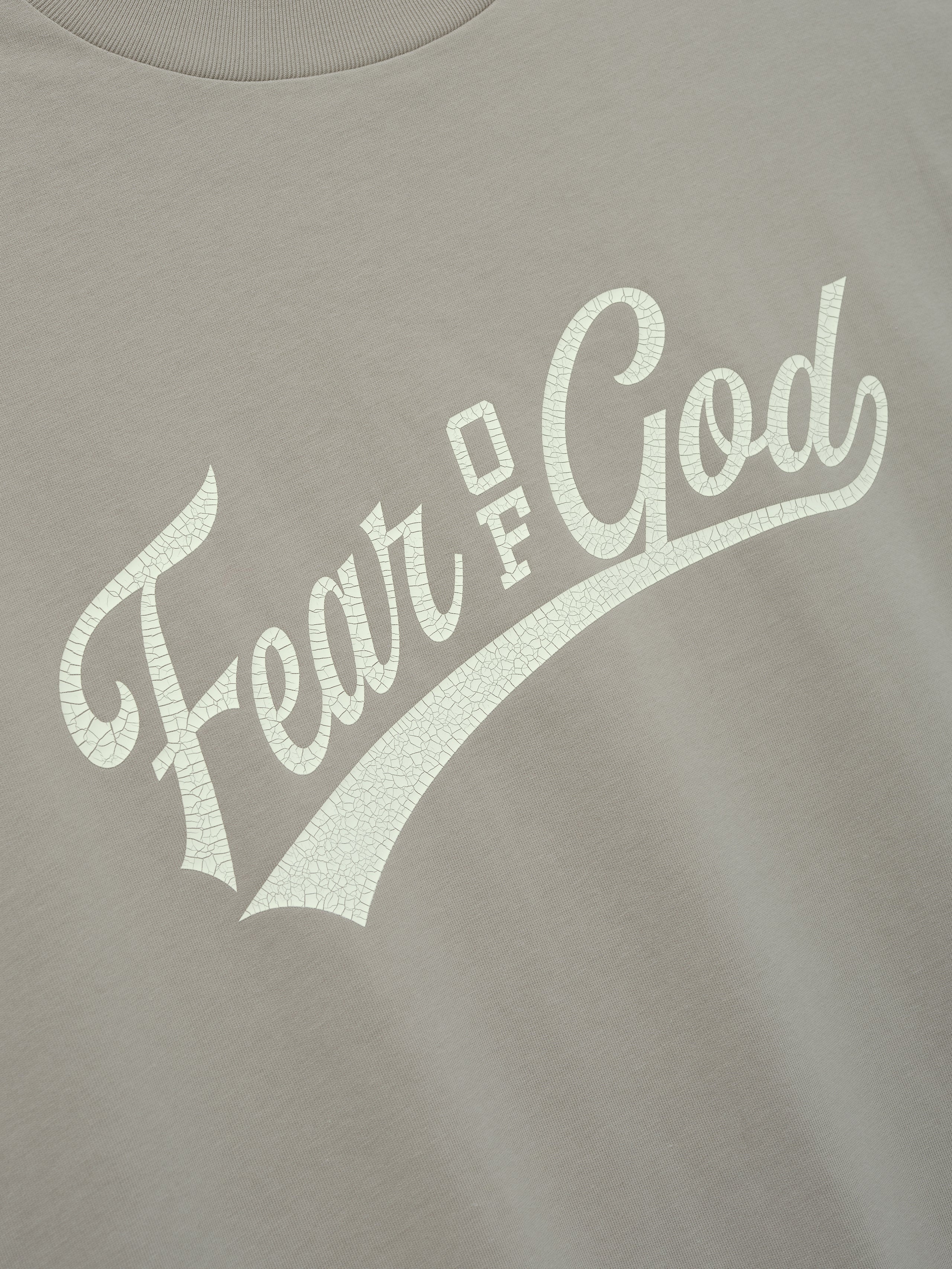 Cut Off 90'S Tee in a muted taupe color featuring a cracked off-white Fear of God script logo across the chest, crafted from soft cotton for a vintage-inspired casual top.