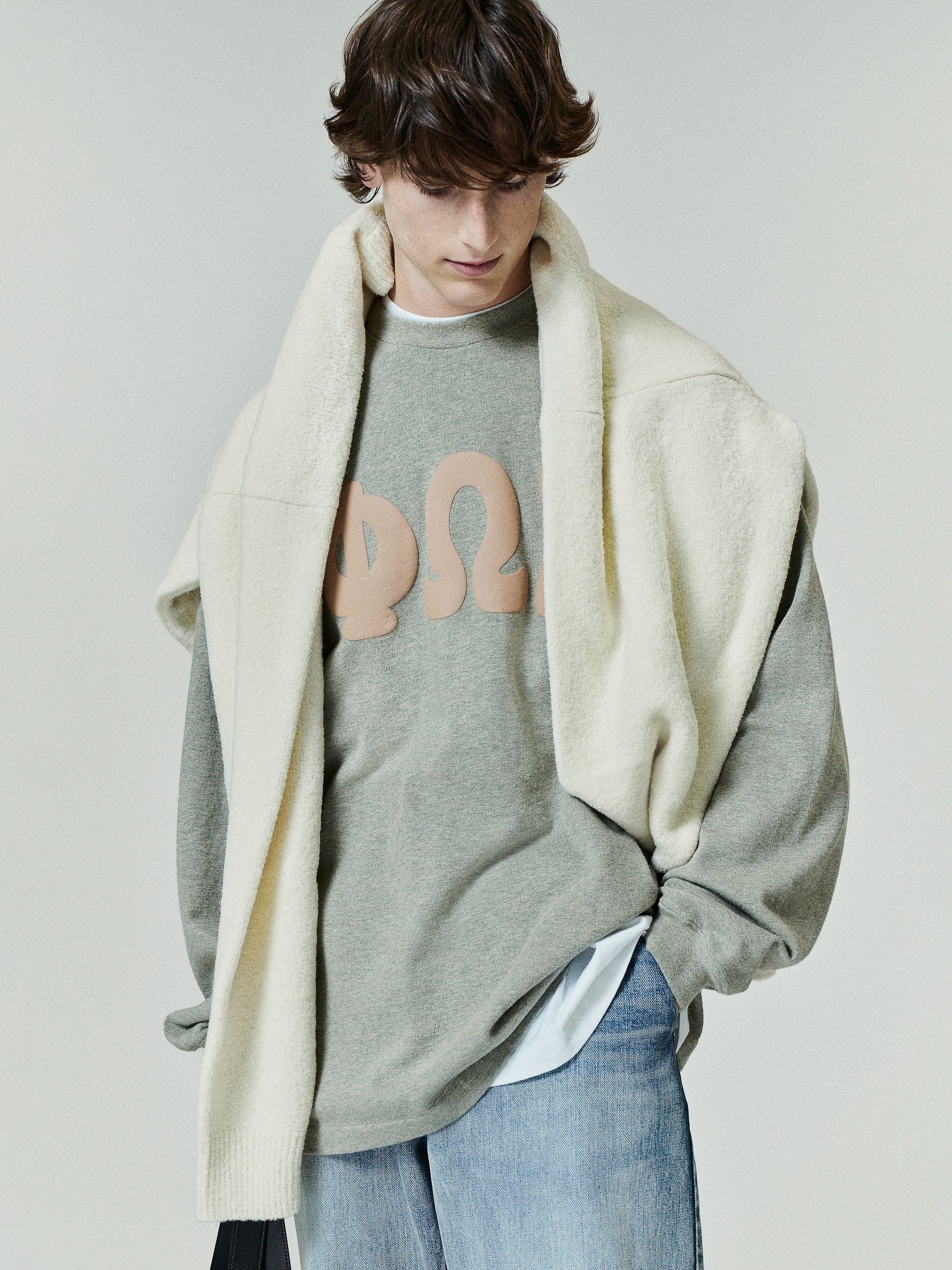 Heavy Jersey Merch Long Sleeve Tee in a soft washed grey-green fabric with large beige Greek letter graphics on the chest, styled with a cream fleece draped over the shoulders for a layered look.