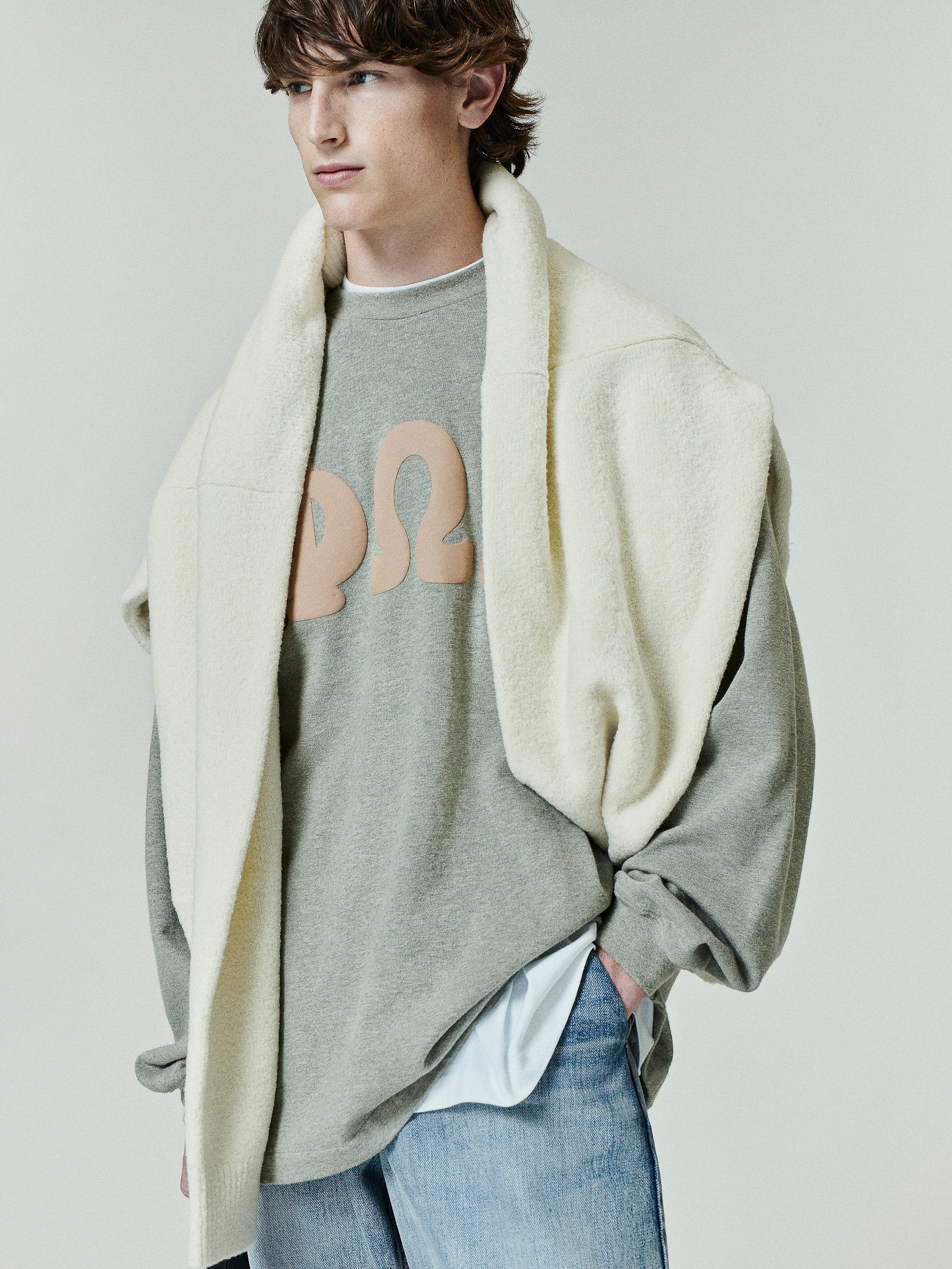Heavy Jersey Merch Long Sleeve Tee in a soft washed grey-green hue featuring large beige Greek letters on the chest, styled with a cream fleece draped over the shoulders for a layered look.