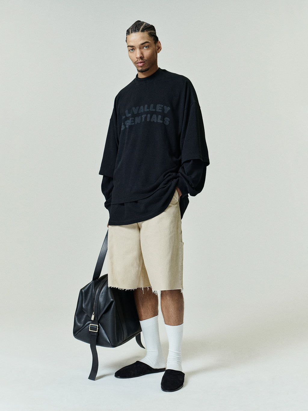 Sport Mesh Merch Tee in vintage black with subtle tonal "ALL VALLEY ESSENTIALS" text, styled with relaxed beige shorts, white crew socks, black slip-on shoes, and a black leather bag.