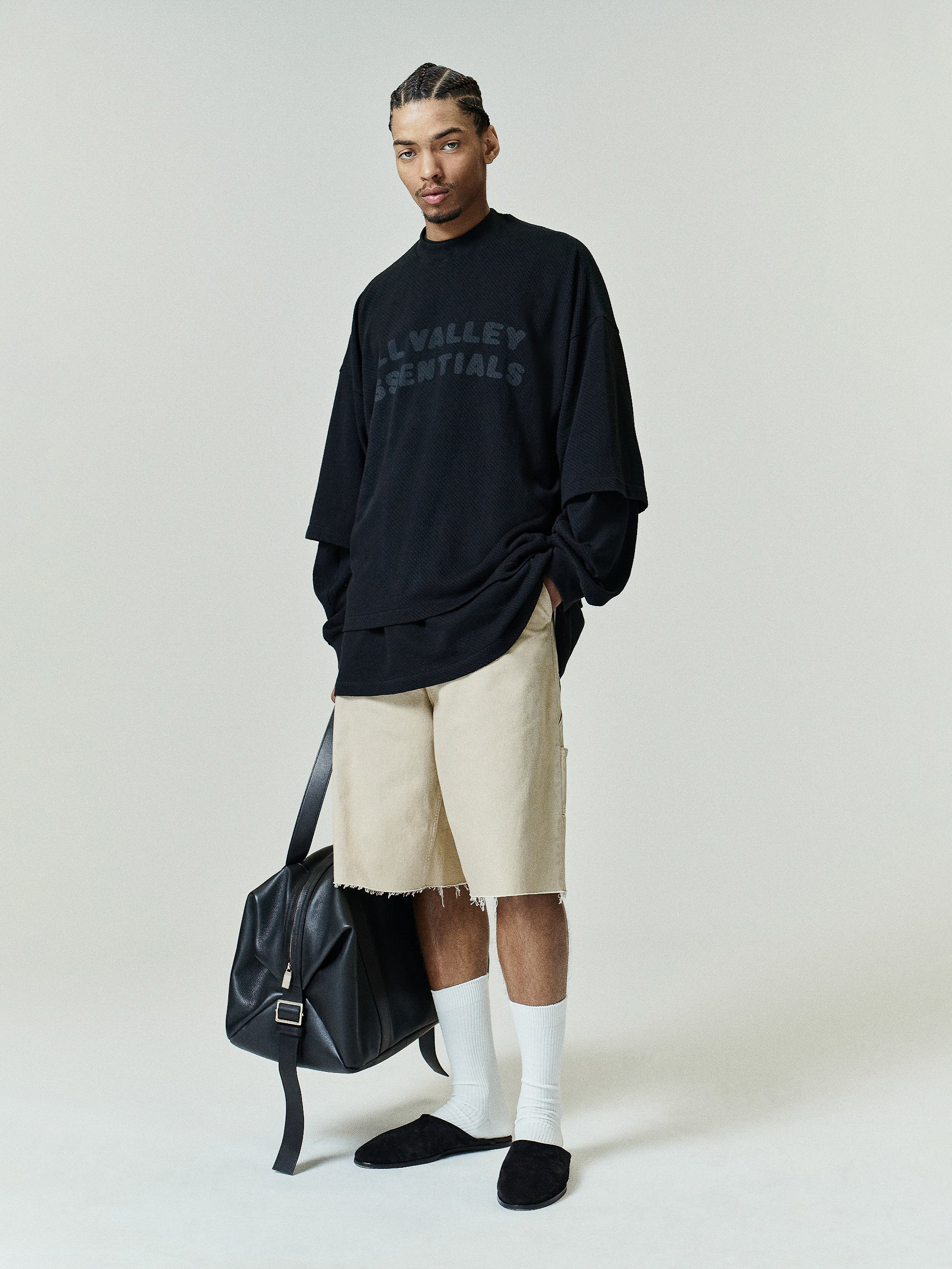 Sport Mesh Merch Tee in vintage black with subtle tonal "ALL VALLEY ESSENTIALS" text, styled with relaxed beige shorts, white crew socks, black slip-on shoes, and a black leather bag.