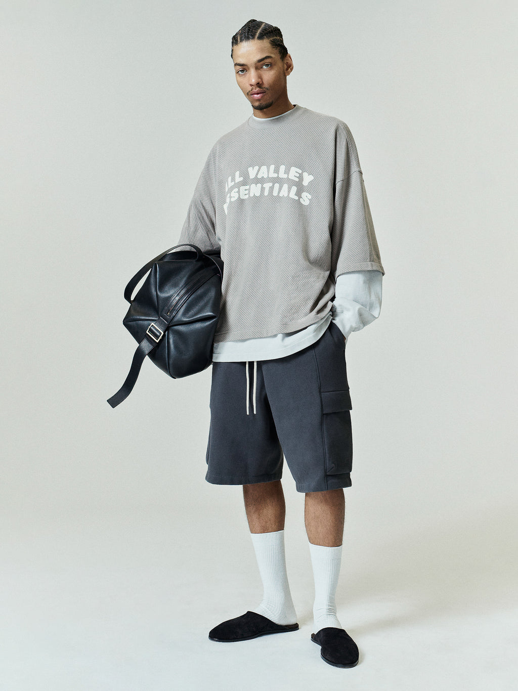 Sport Mesh Merch Tee in a muted seal tone with a relaxed fit, styled with layered long sleeves, dark cargo shorts, white crew socks, and black slip-on shoes, accessorized with a black leather bag.