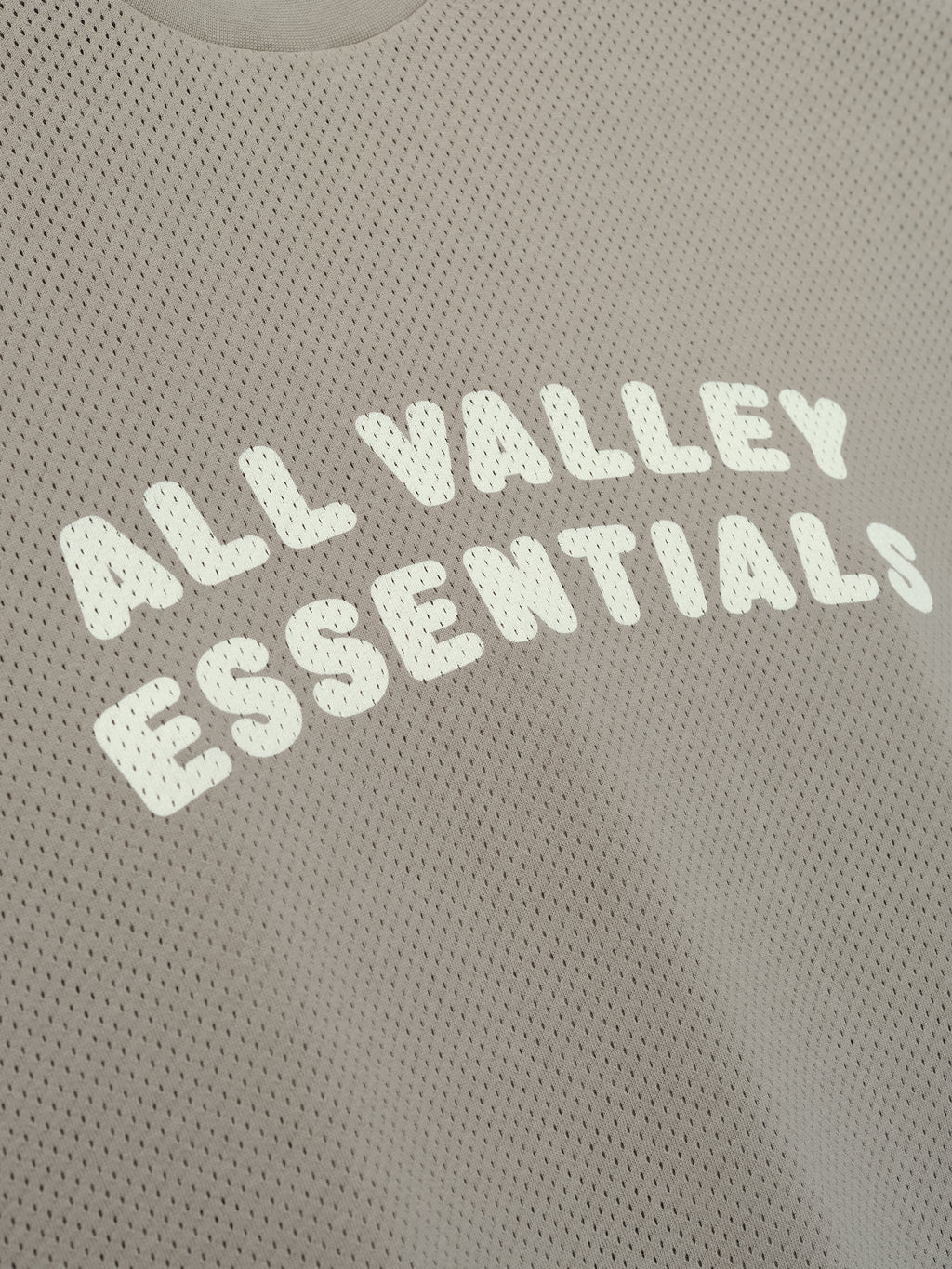 Sport Mesh Merch Tee in a muted taupe shade with breathable mesh fabric, featuring bold white "ALL VALLEY ESSENTIALS" text printed across the chest for a casual, sporty look.