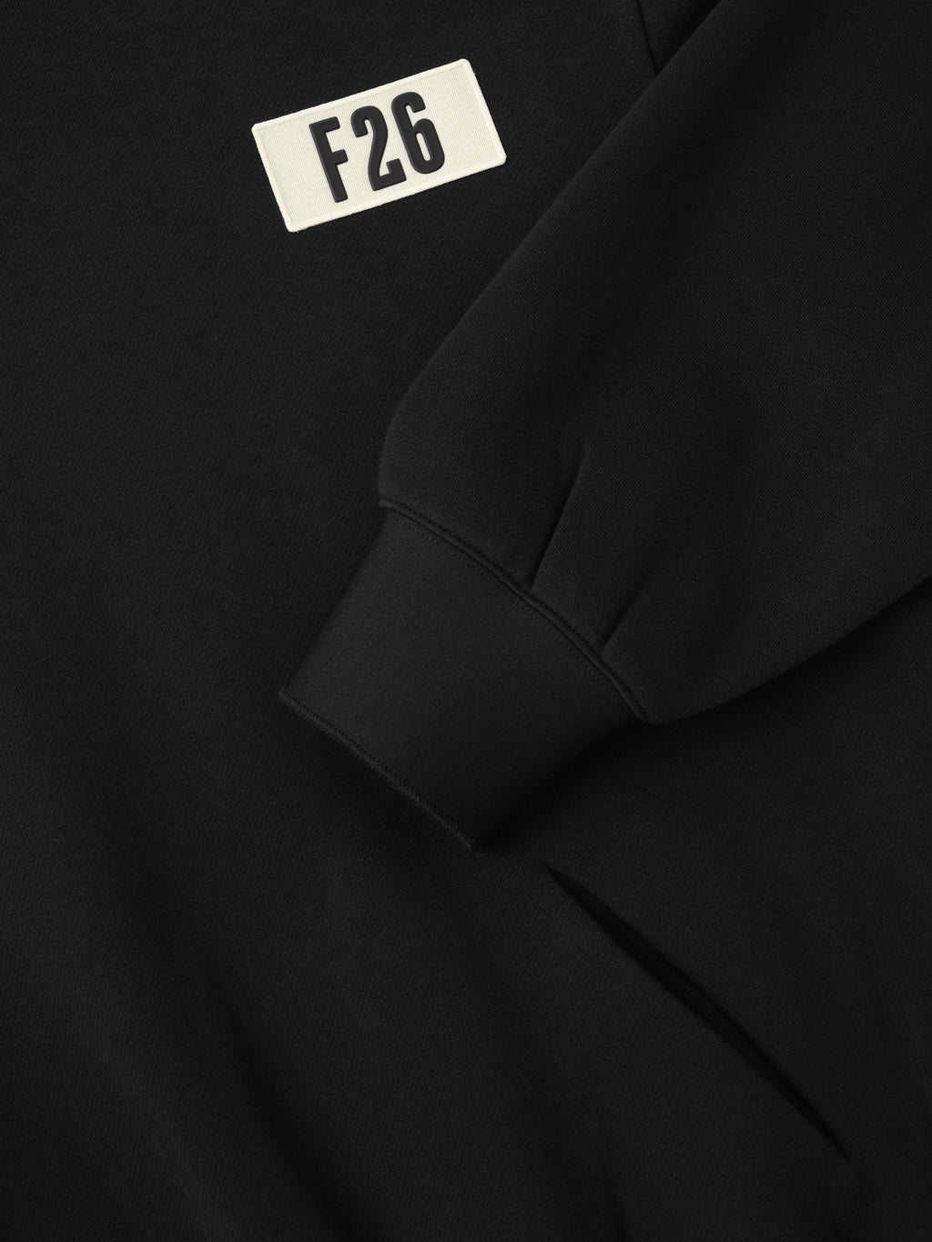 90s Hoodie in deep black fabric featuring a close-up of a ribbed cuff and a small white patch with bold black "F26" lettering on the front chest area.