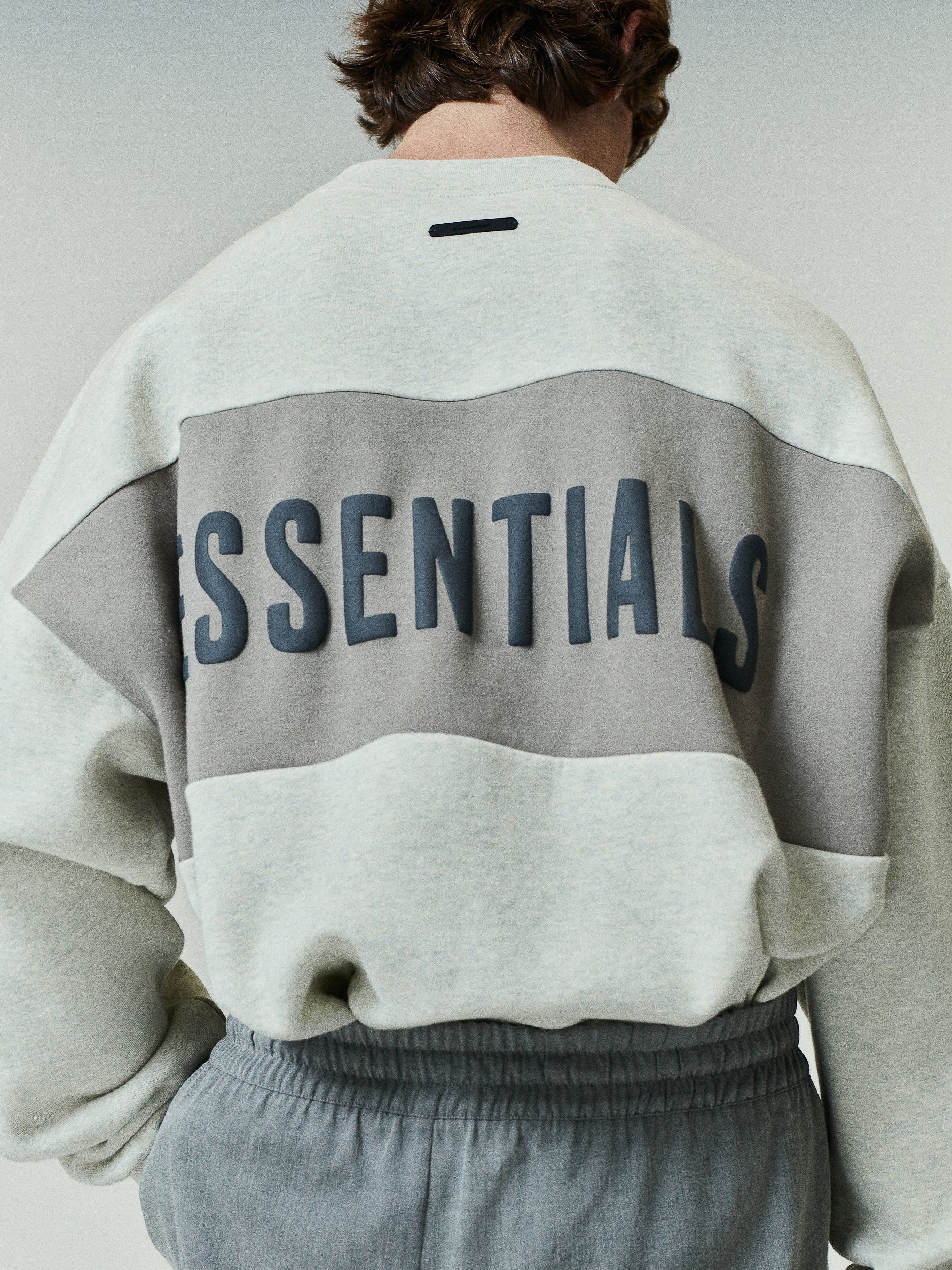 Cropped Relaxed Panelled Sweatshirt in soft vanilla heather with a wide taupe band across the back featuring raised dark ESSENTIALS lettering for a refined streetwear appeal.