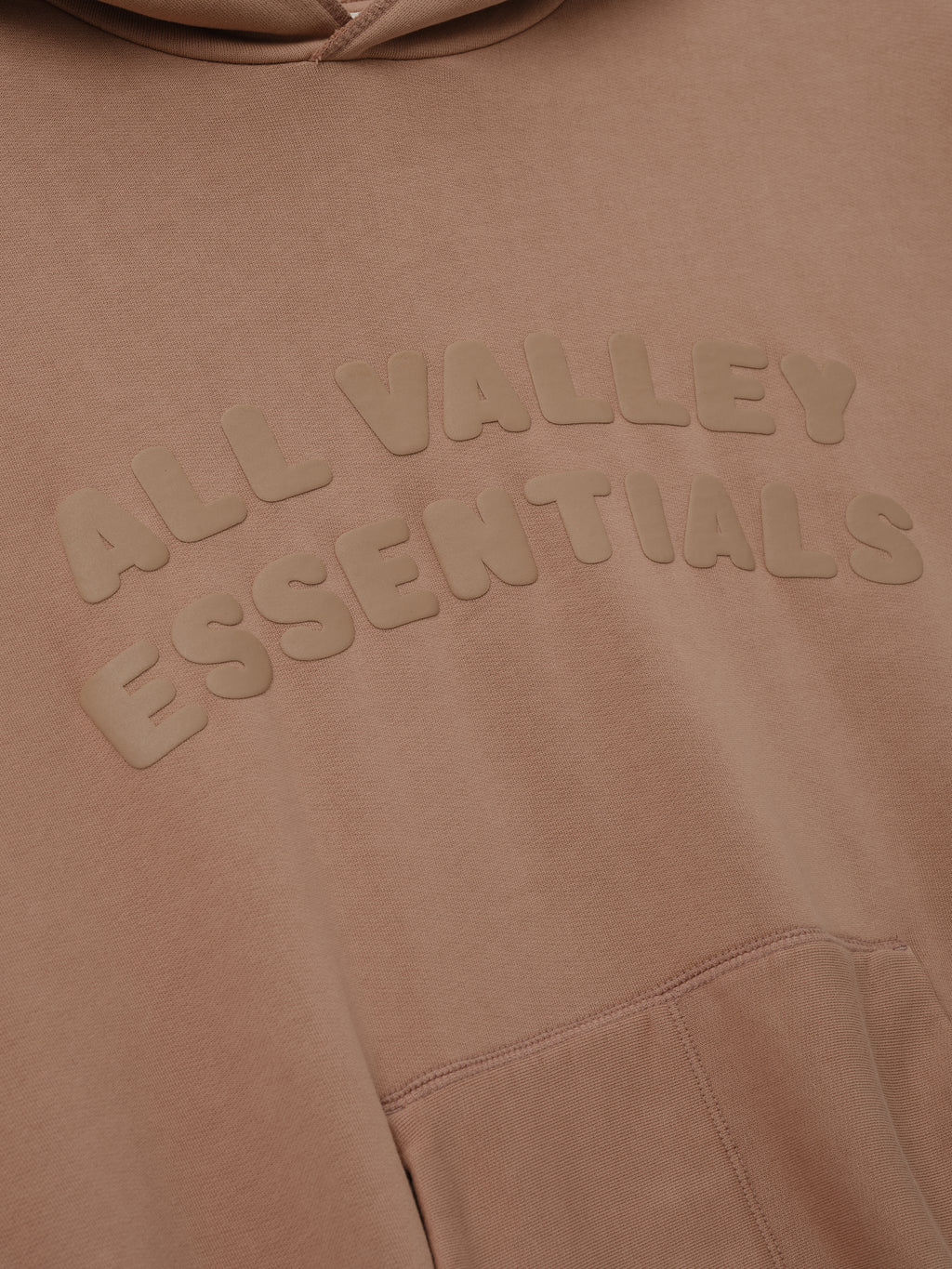 90s Hoodie in washed ginger tone featuring a close-up of raised tonal lettering "ALL VALLEY ESSENTIALS" across the chest with a kangaroo pocket seam visible below.