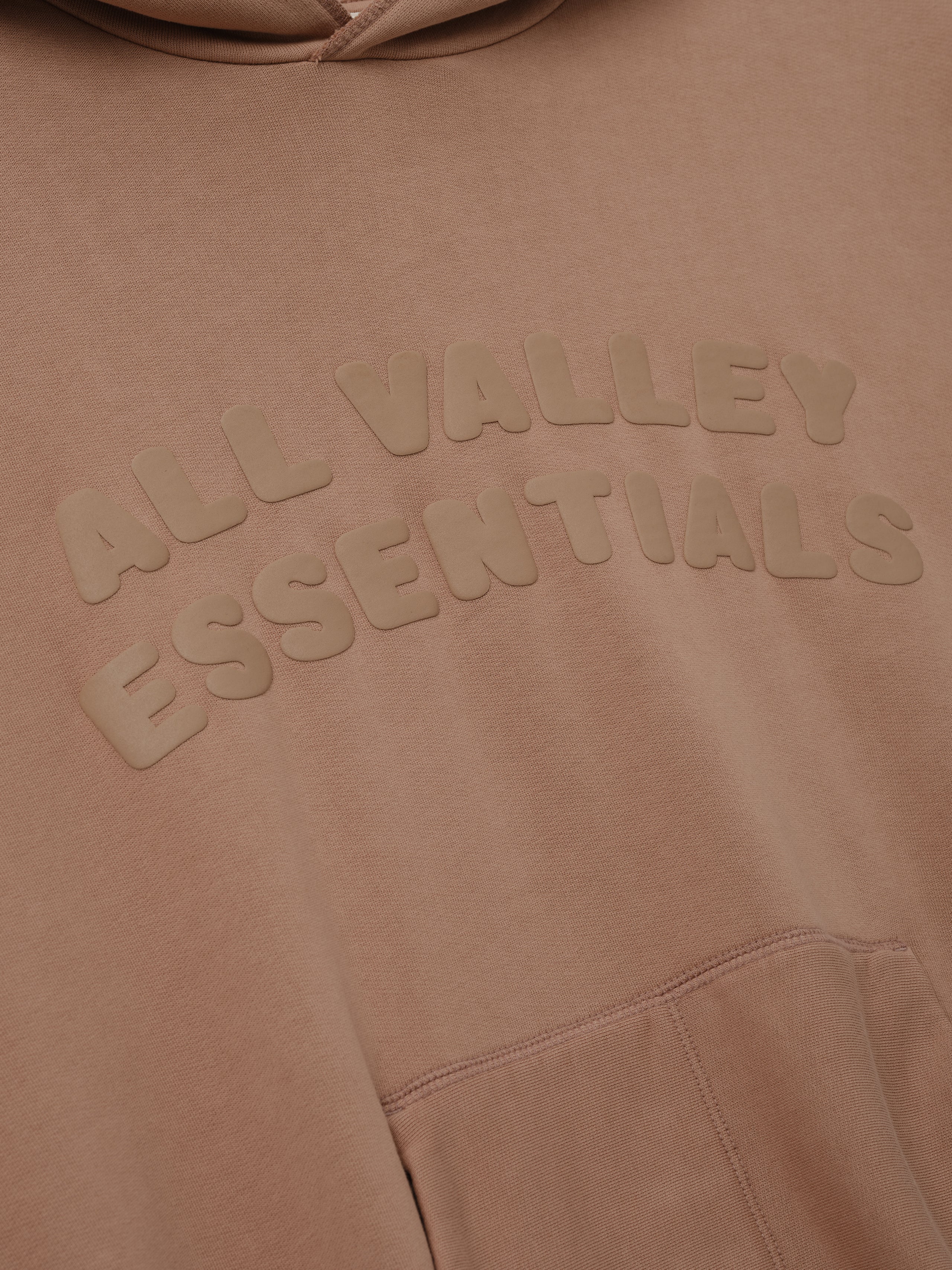90s Hoodie in washed ginger tone featuring a close-up of raised tonal lettering "ALL VALLEY ESSENTIALS" across the chest with a kangaroo pocket seam visible below.