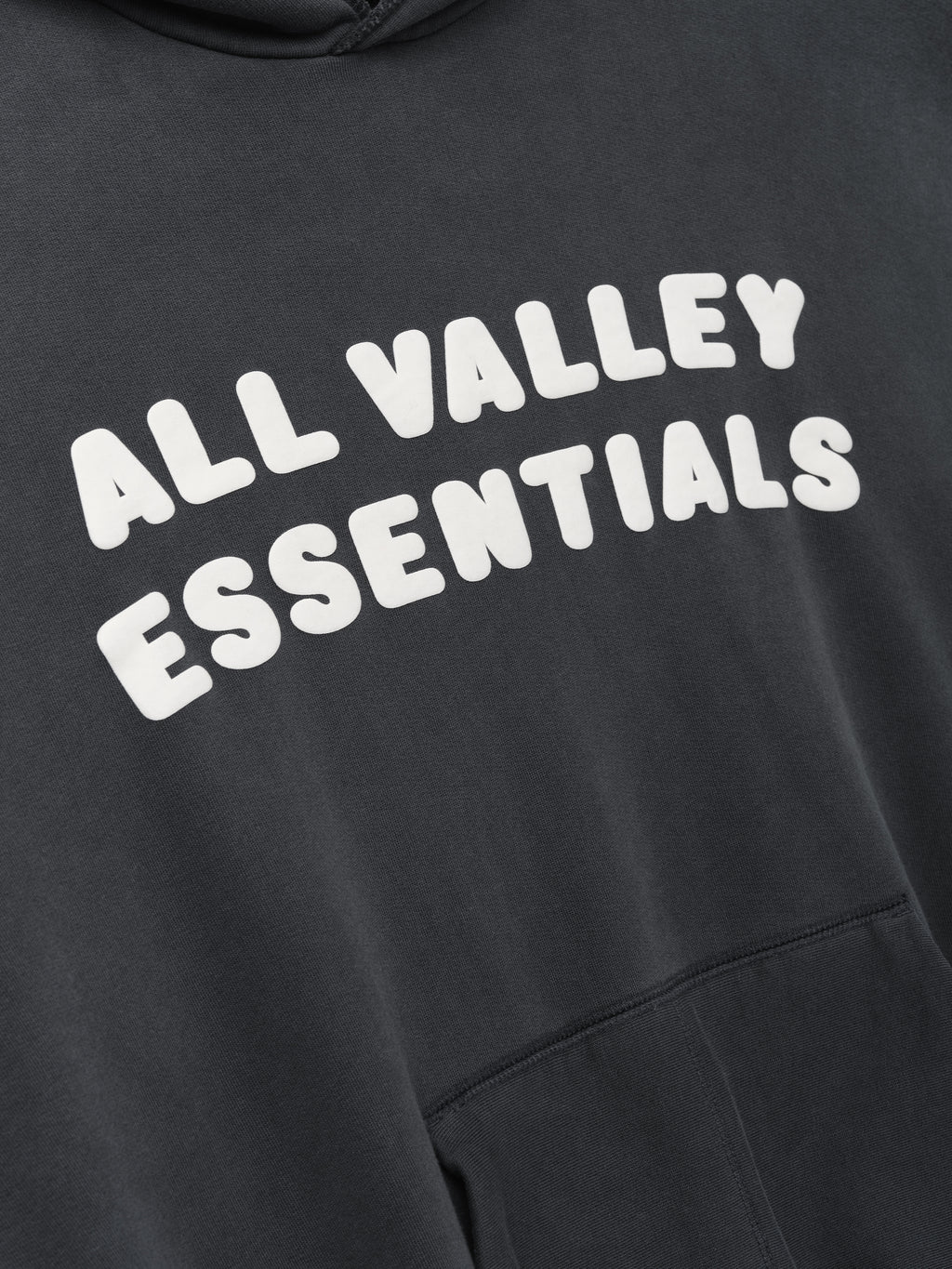 90s Hoodie in faded iron grey showing a close-up of the chest with bold white raised lettering "ALL VALLEY ESSENTIALS" and part of the front kangaroo pocket on soft fabric.