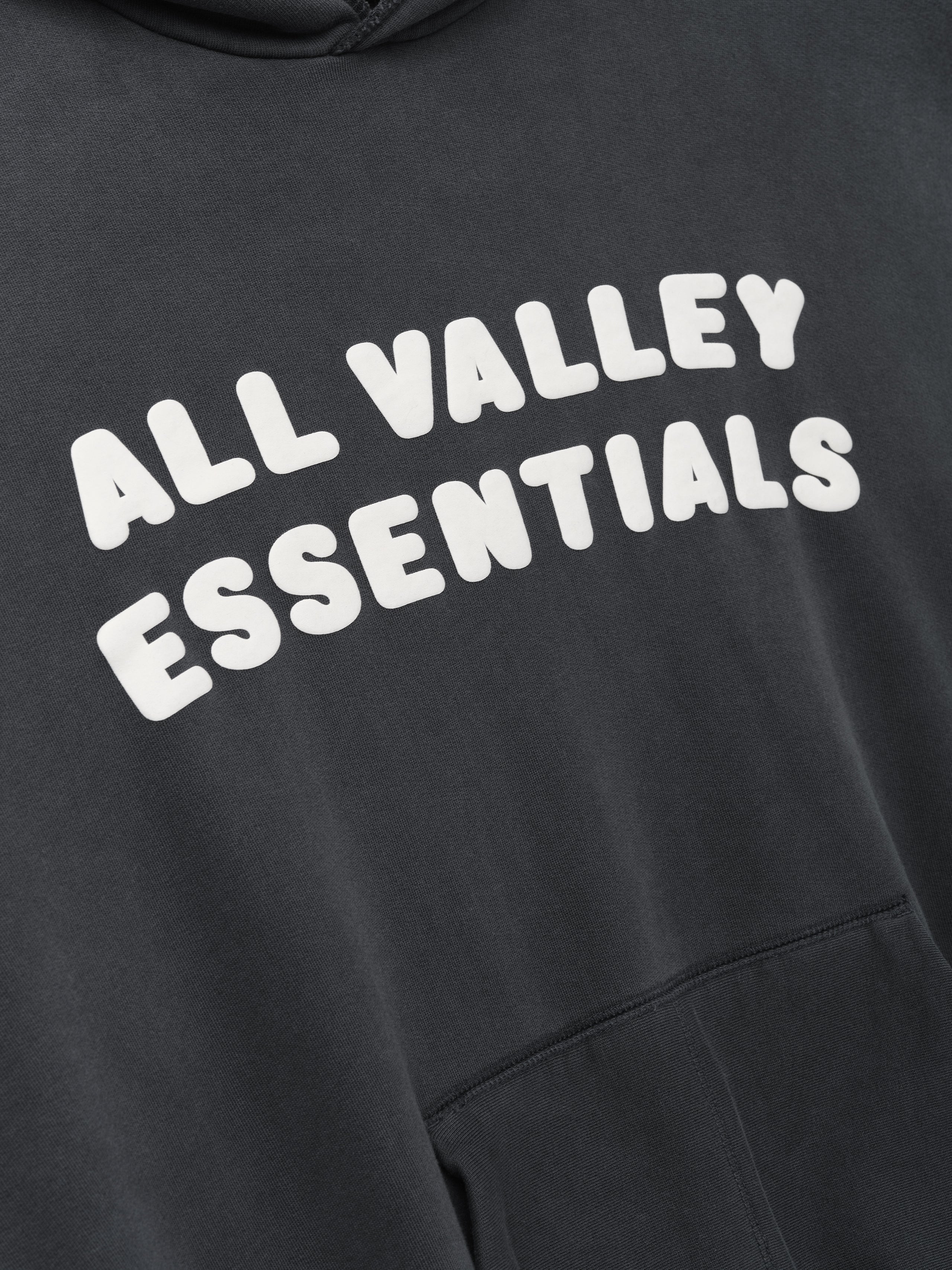 90s Hoodie in faded iron grey showing a close-up of the chest with bold white raised lettering "ALL VALLEY ESSENTIALS" and part of the front kangaroo pocket on soft fabric.