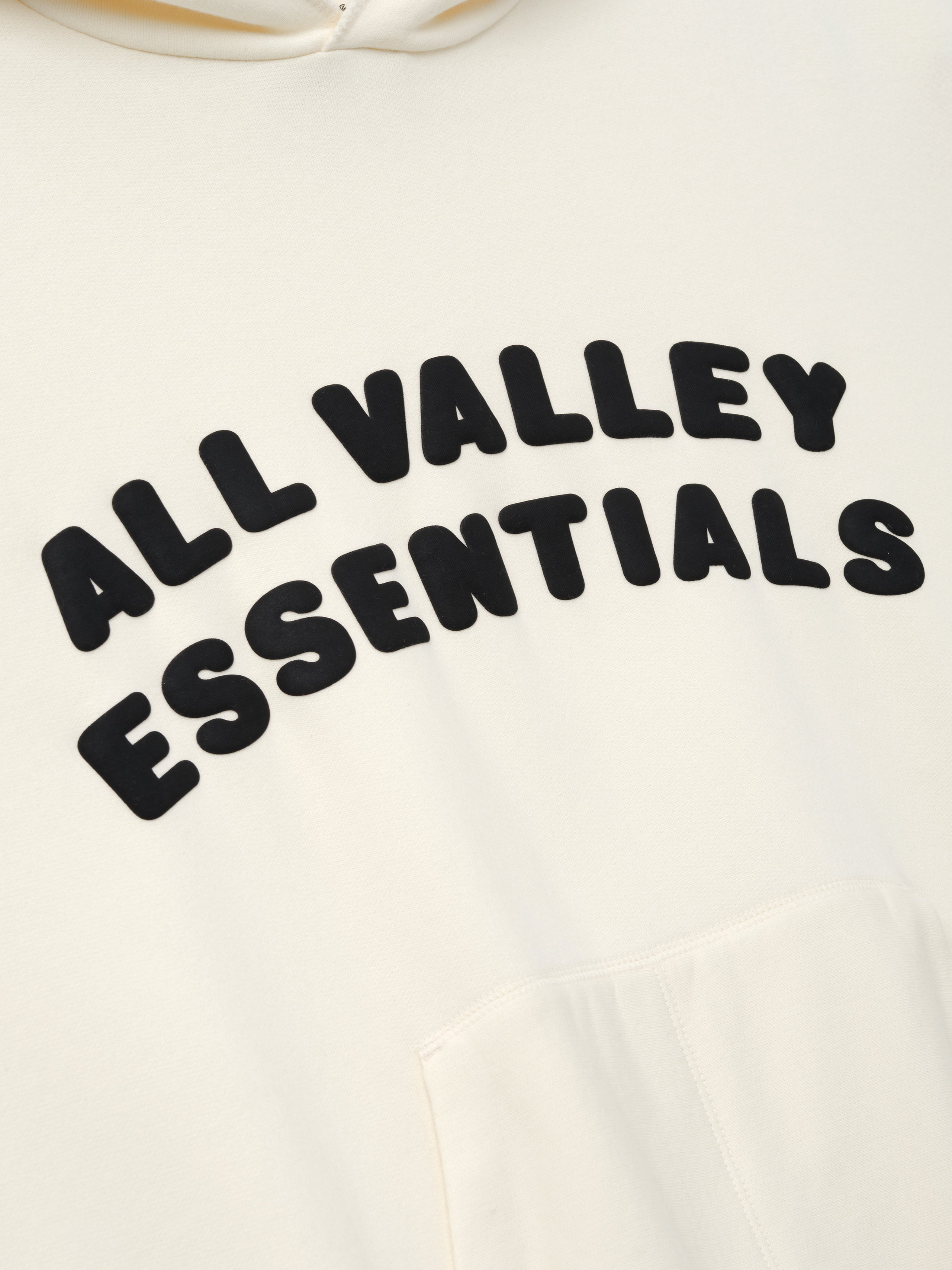 90s Hoodie in a soft washed ivory tone featuring raised black lettering "ALL VALLEY ESSENTIALS" arched across the chest and a roomy kangaroo pocket below on cozy outerwear.