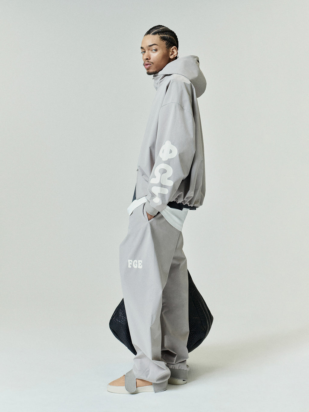 Model wearing a light gray Storm Pant with a relaxed fit, featuring side pockets and subtle branding near the thigh, paired with a matching oversized hooded jacket and neutral-toned footwear.