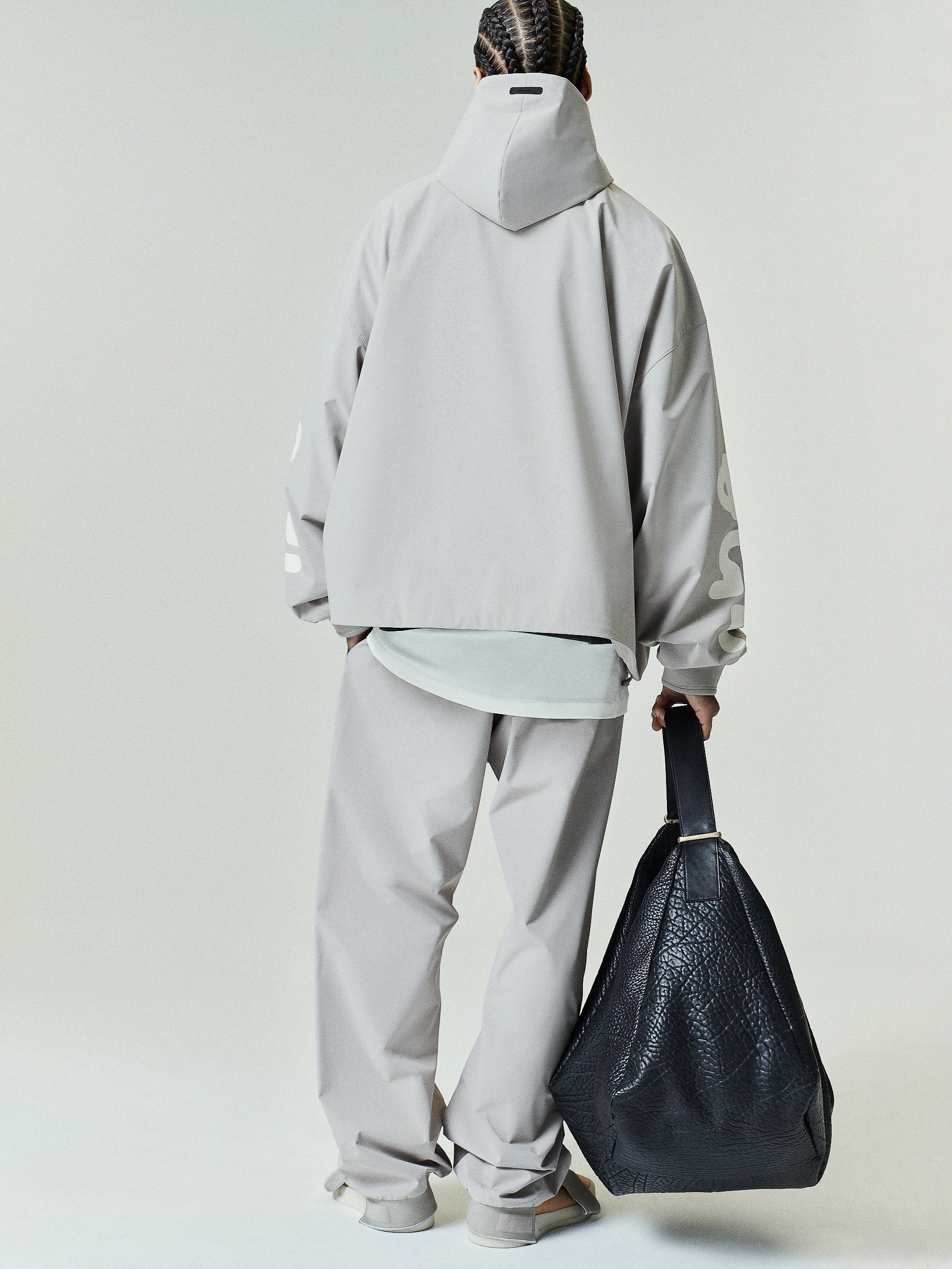 Model wearing a loose light gray Storm Pant paired with a matching oversized hooded jacket, holding a large black textured leather tote bag, viewed from behind.