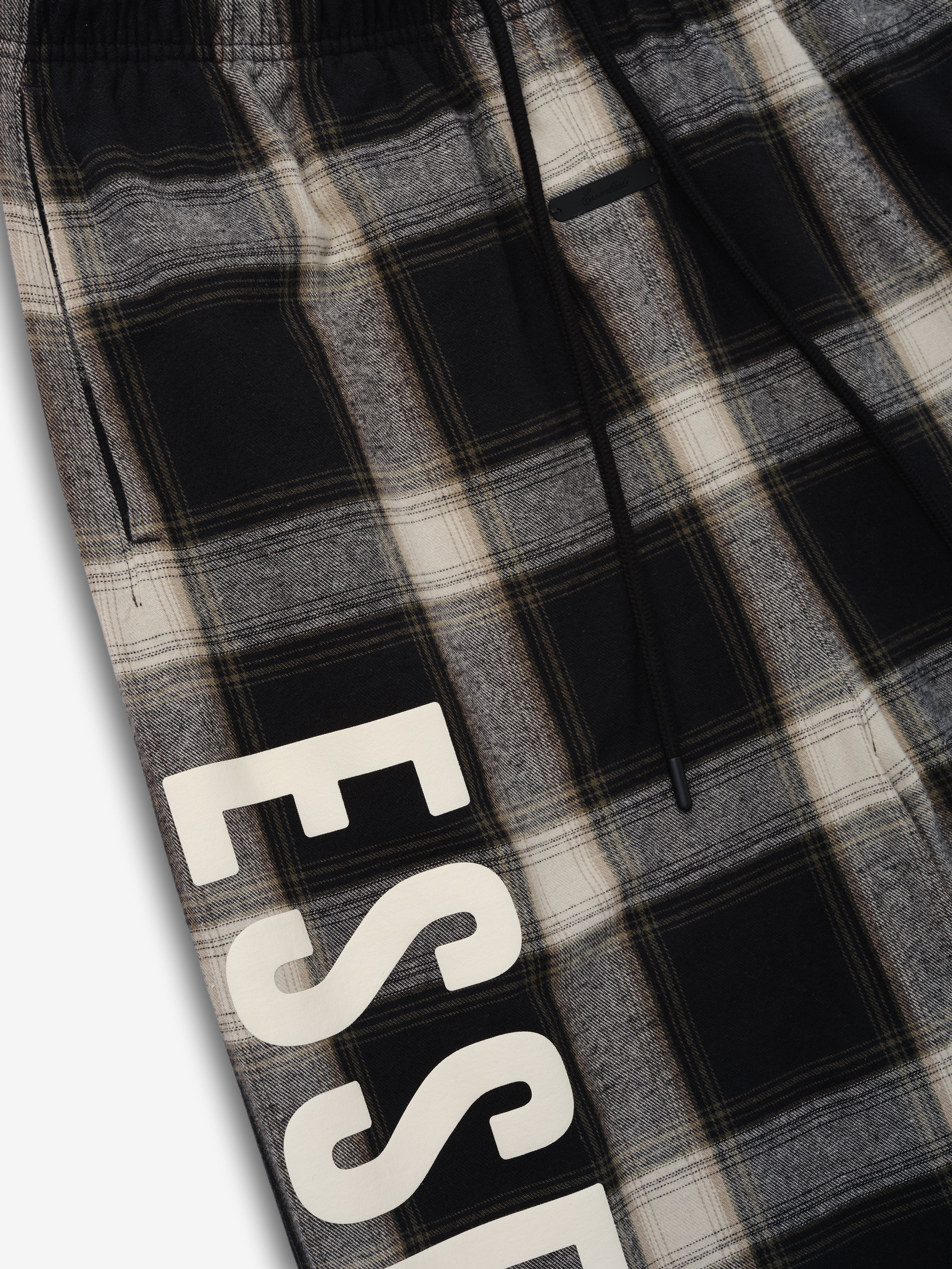 Flannel Lounge Pant in a muted gray and black plaid pattern, featuring an elastic waistband, side pockets, and large cream-colored "ESSENTIALS" lettering along the leg for a modern casual look.