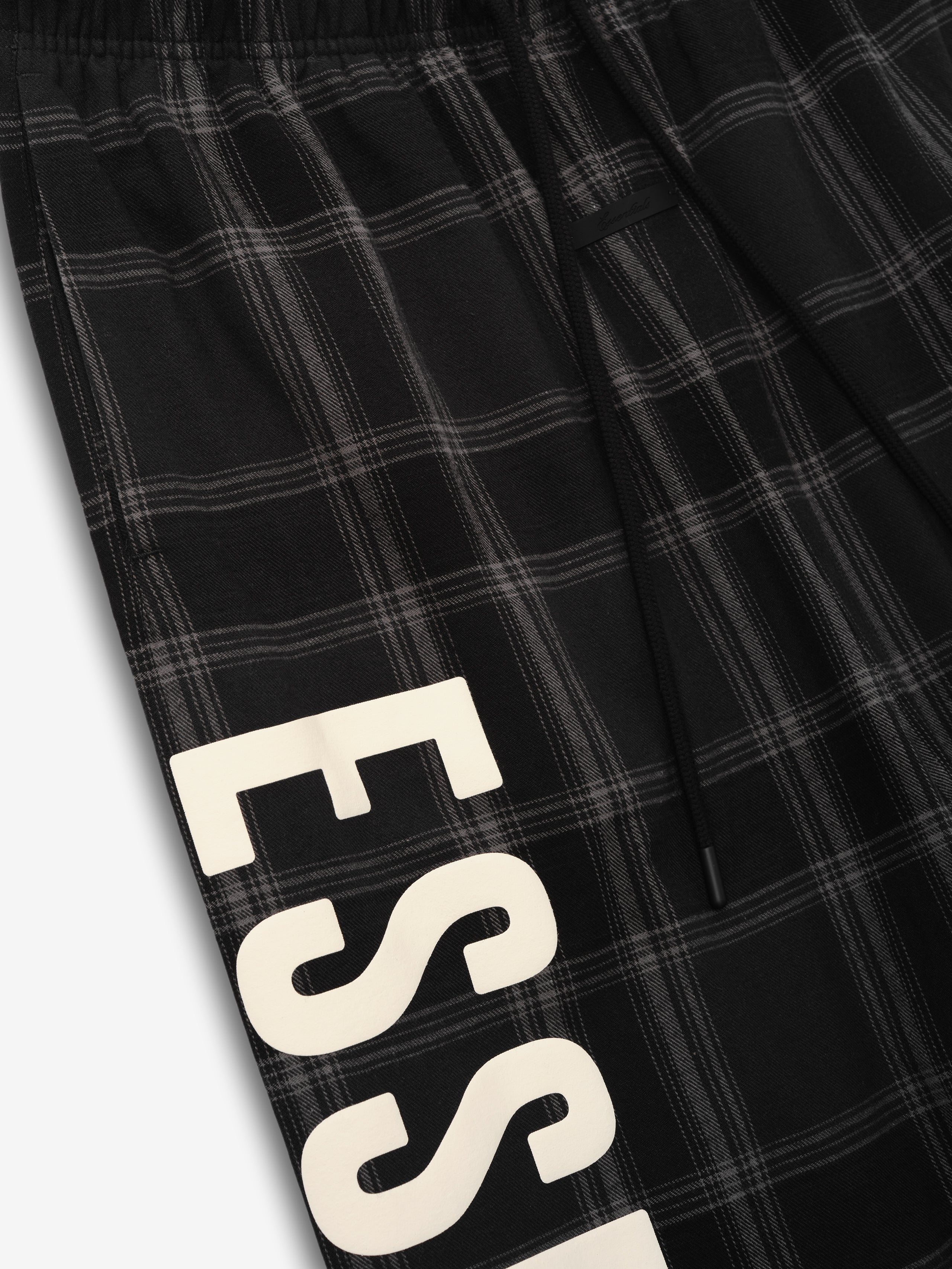 Flannel Lounge Pant featuring a black and gray plaid pattern with an elastic waistband and a partial bold cream-colored vertical "ESSENTIALS" print along the leg.