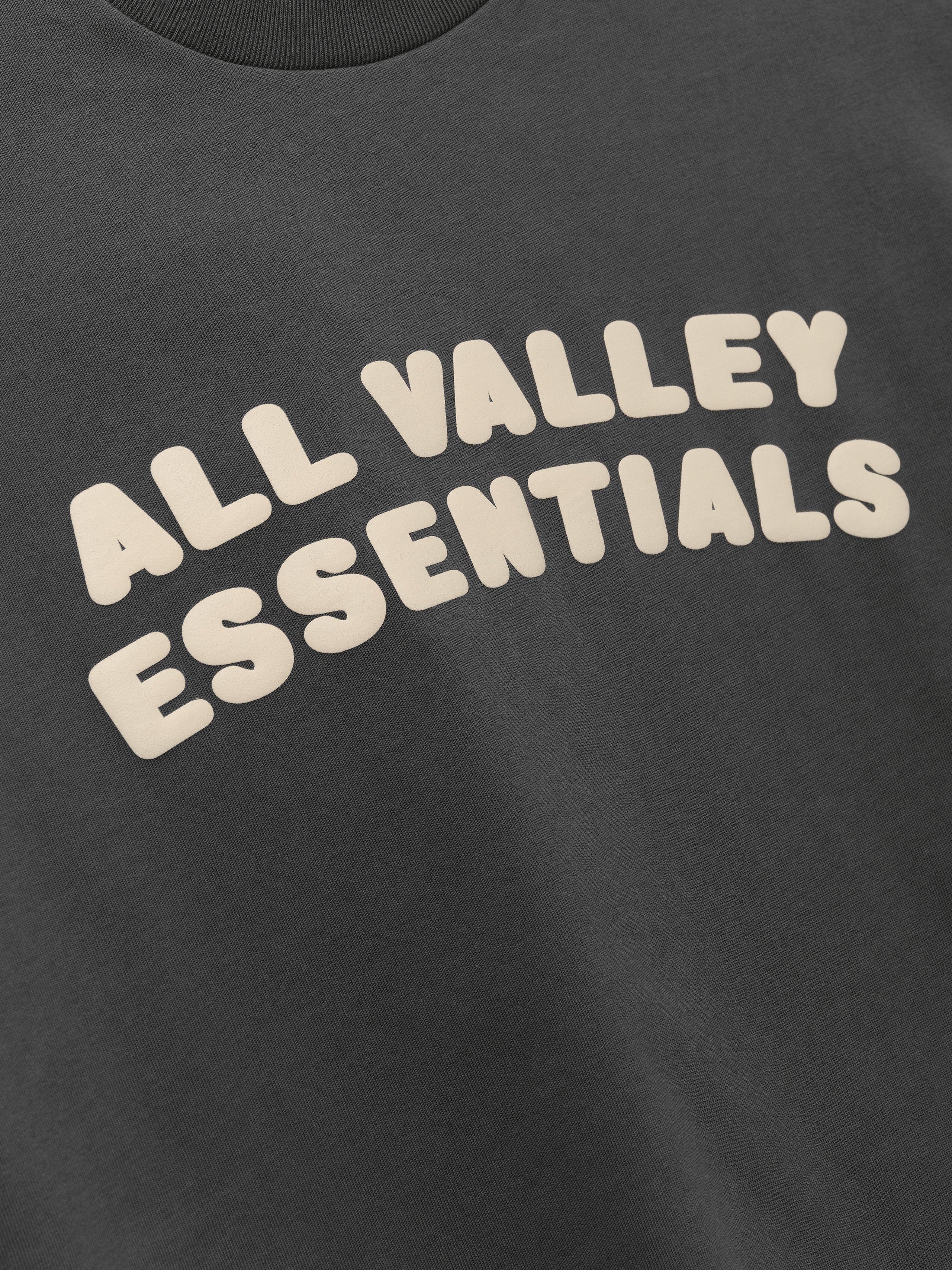 Kids Merch Tee in iron gray fabric featuring playful cream-colored bubble text "ALL VALLEY ESSENTIALS" across the front for a fun, casual kids' top.