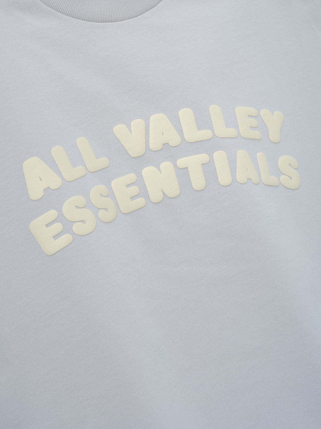 Kids Merch Tee in pale gray fabric featuring soft cream-colored raised text "ALL VALLEY ESSENTIALS" arranged in a curved design for a comfortable casual kids' top.