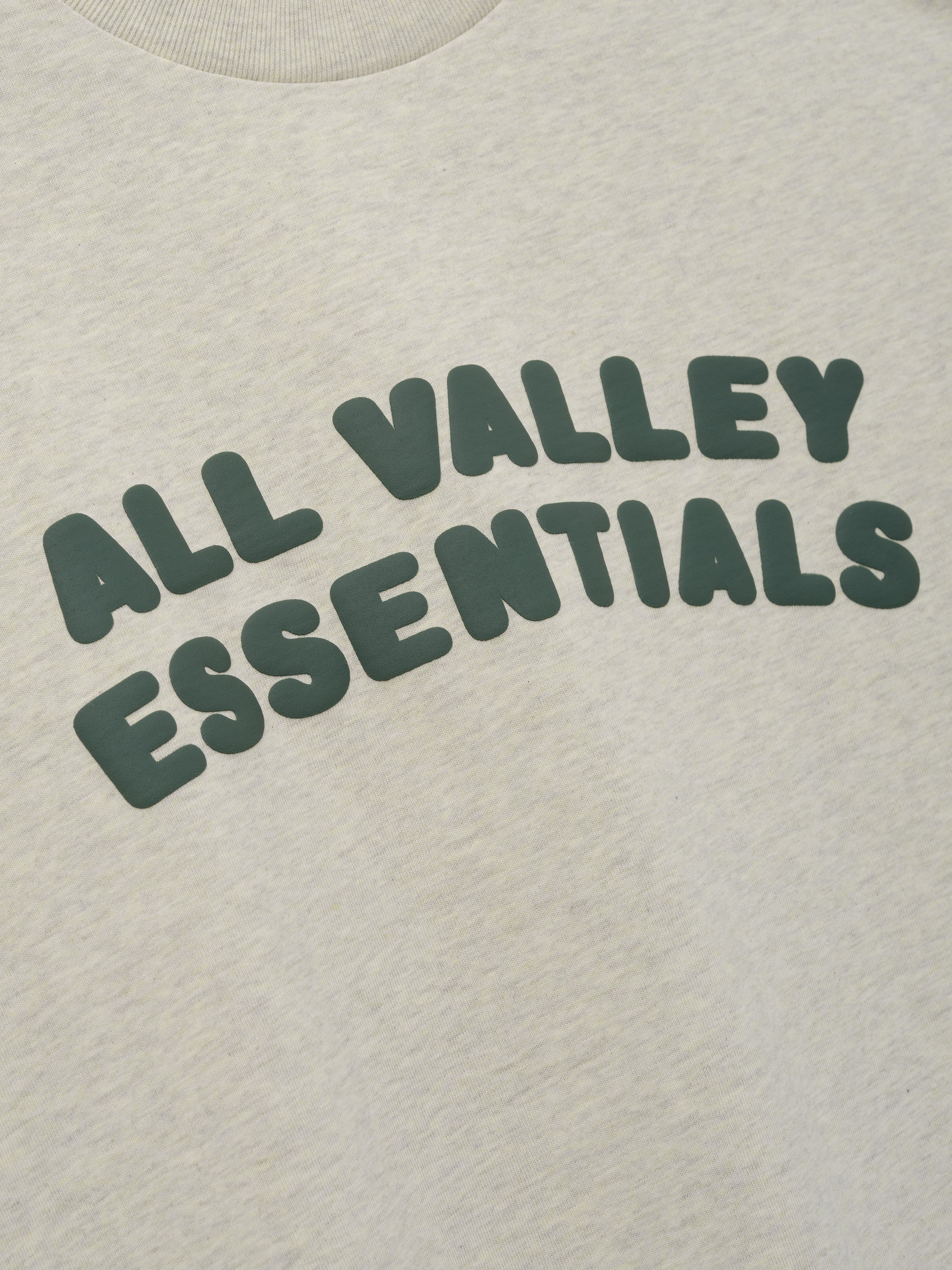 Kids Merch Tee in soft vanilla heather fabric featuring dark green rounded bubble lettering "ALL VALLEY ESSENTIALS" in a relaxed arch, designed for casual kids’ tops.