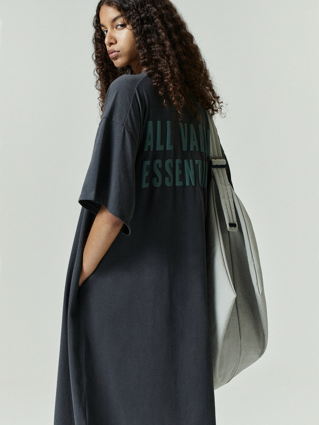 Model wears Womens Long TShirt Dress in dark gray with oversized fit and short sleeves, featuring subtle dark green lettering on the back and paired with a large light gray tote bag.