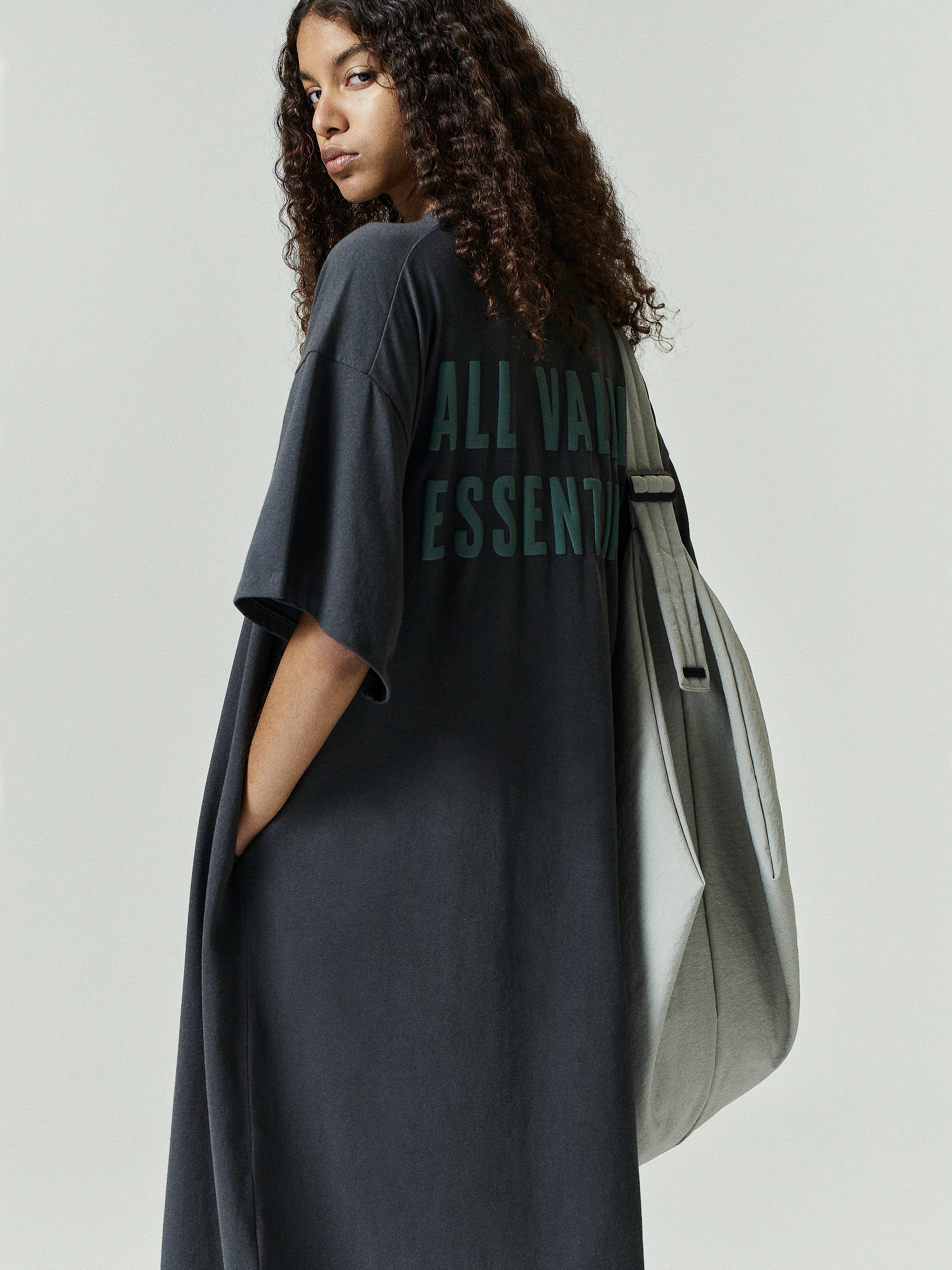 Model wears Womens Long TShirt Dress in dark gray with oversized fit and short sleeves, featuring subtle dark green lettering on the back and paired with a large light gray tote bag.