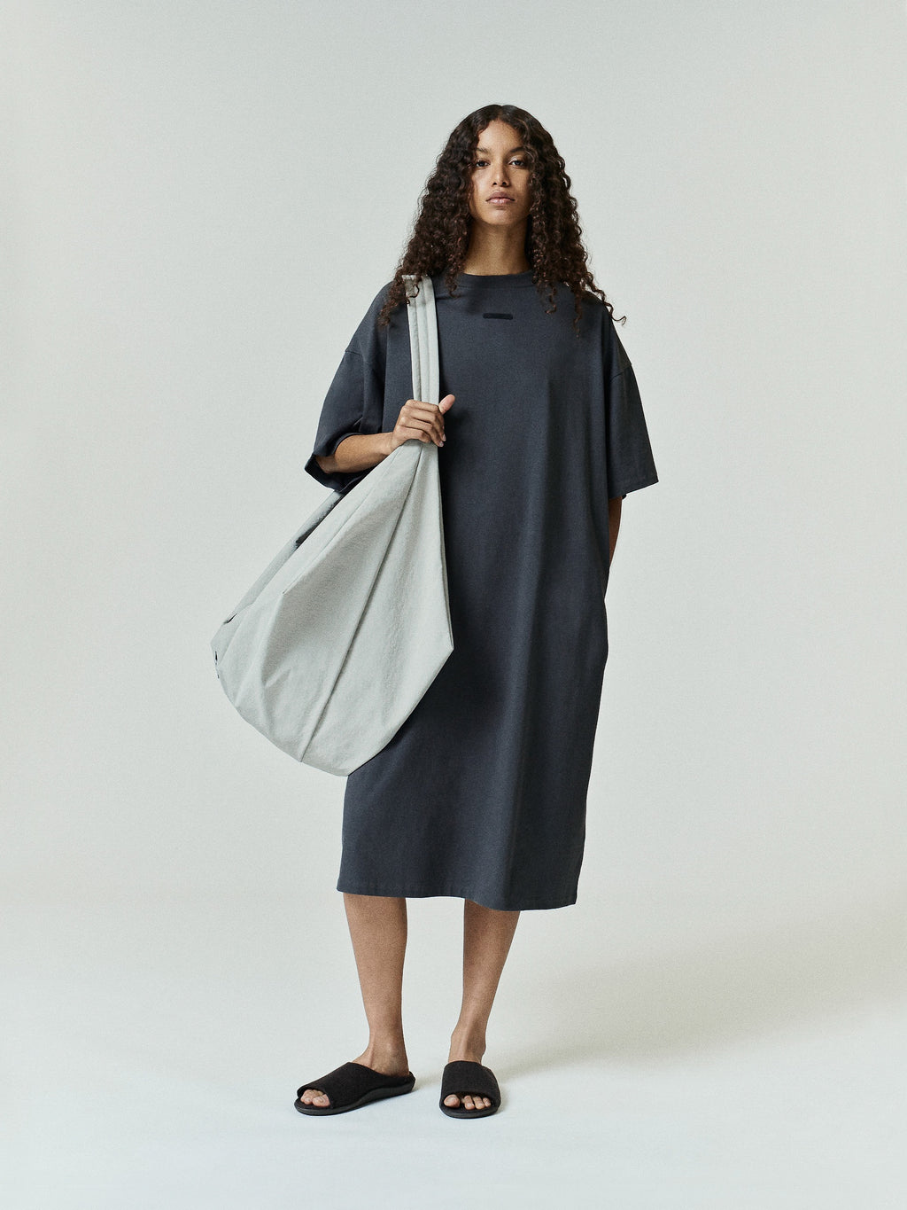 Model wearing Womens Long TShirt Dress in deep charcoal gray with a relaxed, oversized silhouette and short sleeves, paired with a large soft gray tote bag and black slide sandals.