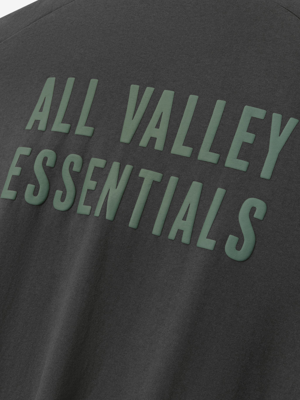 Close-up of dark gray Womens Long TShirt Dress fabric featuring raised green lettering reading "ALL VALLEY ESSENTIALS" across the back in bold, capital letters.