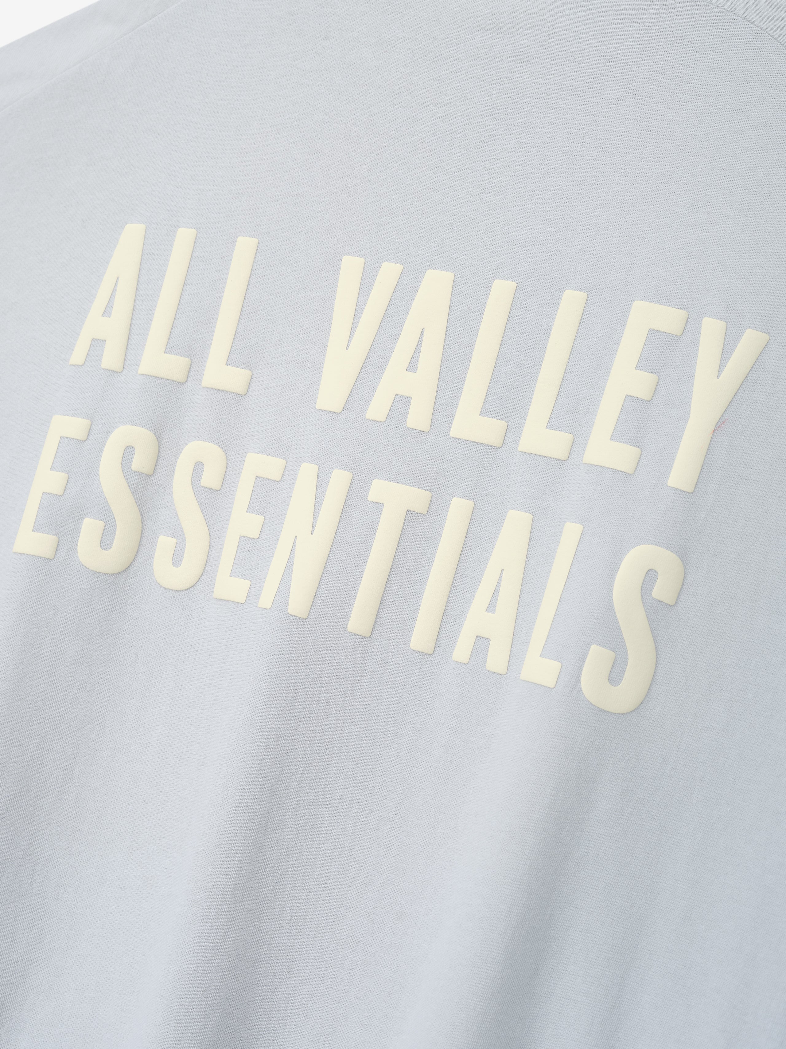 Close-up of Womens Long TShirt Dress fabric in soft light gray featuring raised cream-colored lettering reading "ALL VALLEY ESSENTIALS" in a clean, bold font.