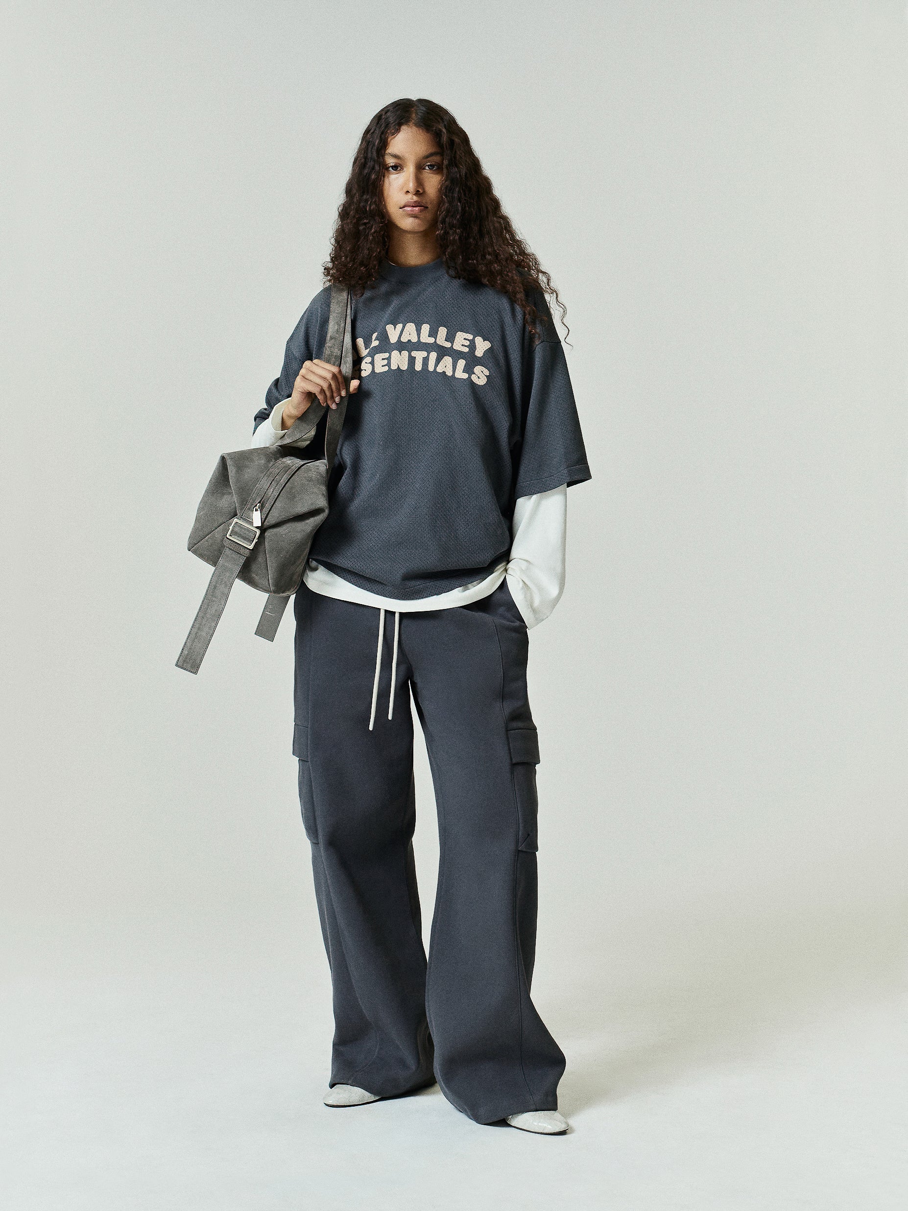 Womens Sport Mesh Merch Tee in muted dark grey with bold textured lettering across the chest, styled layered over a long-sleeve top paired with wide-leg pants for an effortlessly cool look.