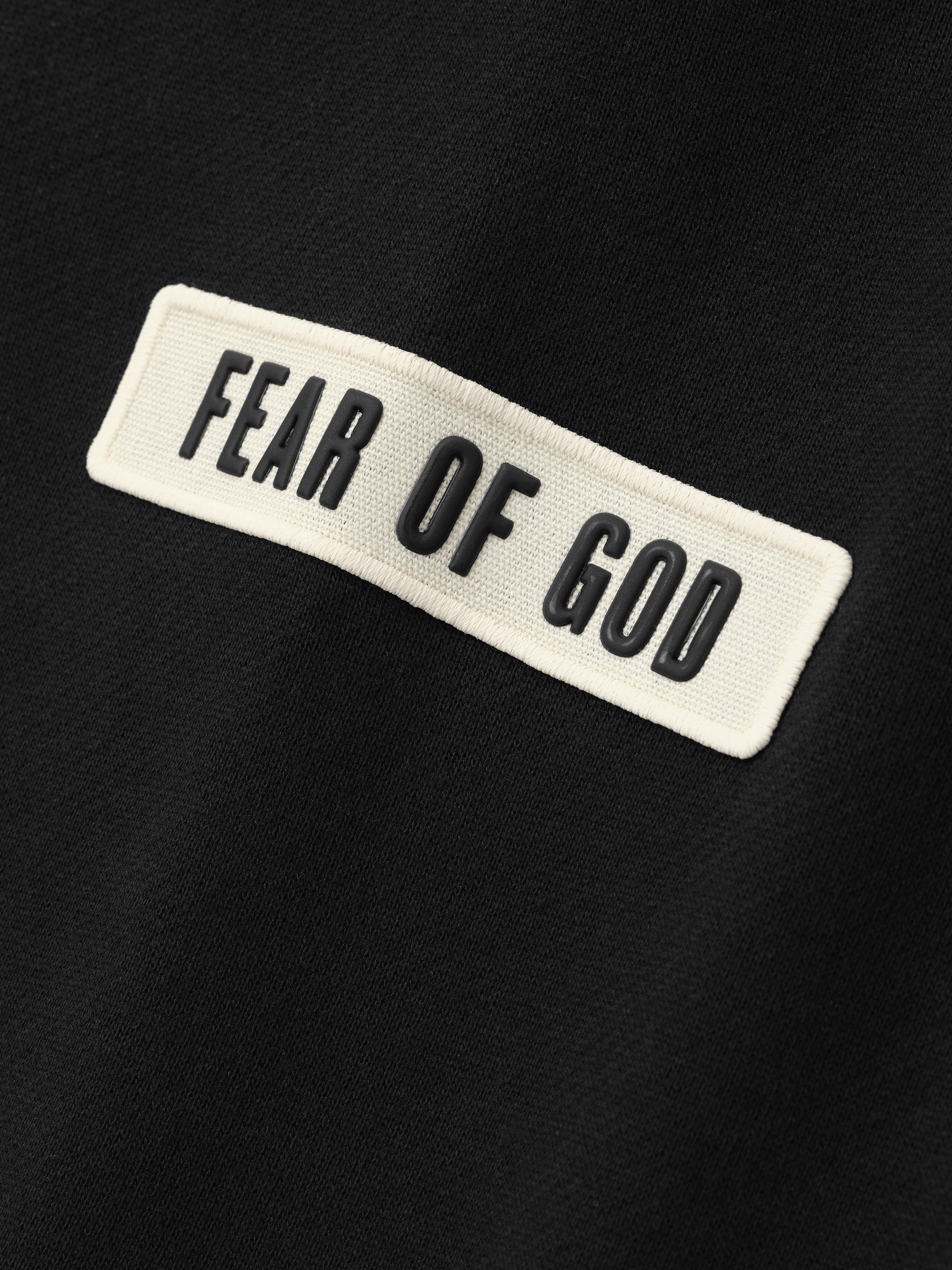 Close-up of a soft black fabric featuring a contrasting white rectangular patch with raised black lettering that reads "Fear of God" on a Classic Fit Sweatpant.